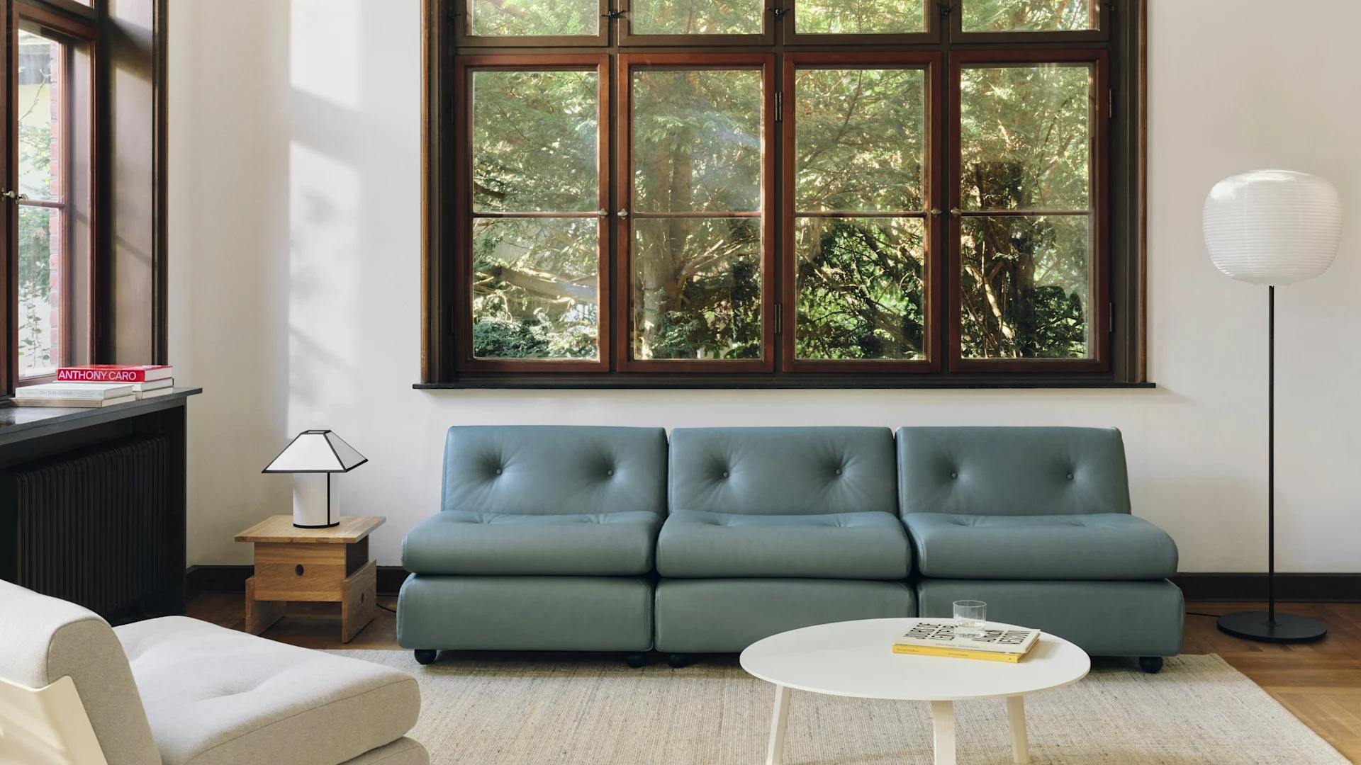 Salon moderne avec un canapé modulaire bleu-gris, une table basse blanche et une grande fenêtre donnant sur des arbres verts. Deux lampes éclairent la pièce.