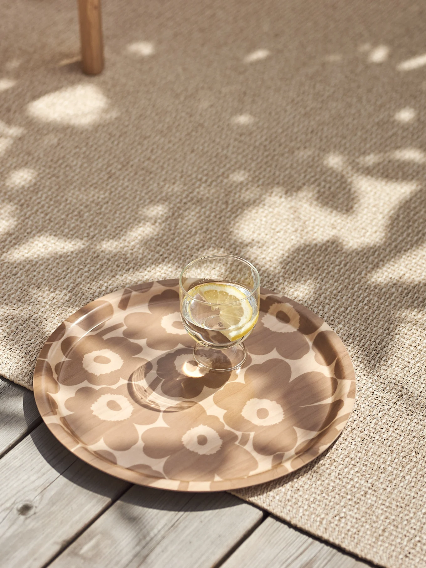 Verre d'eau citronnée sur un plateau beige à motif floral posé sur un tapis texturé avec de la lumière solaire tachetée.