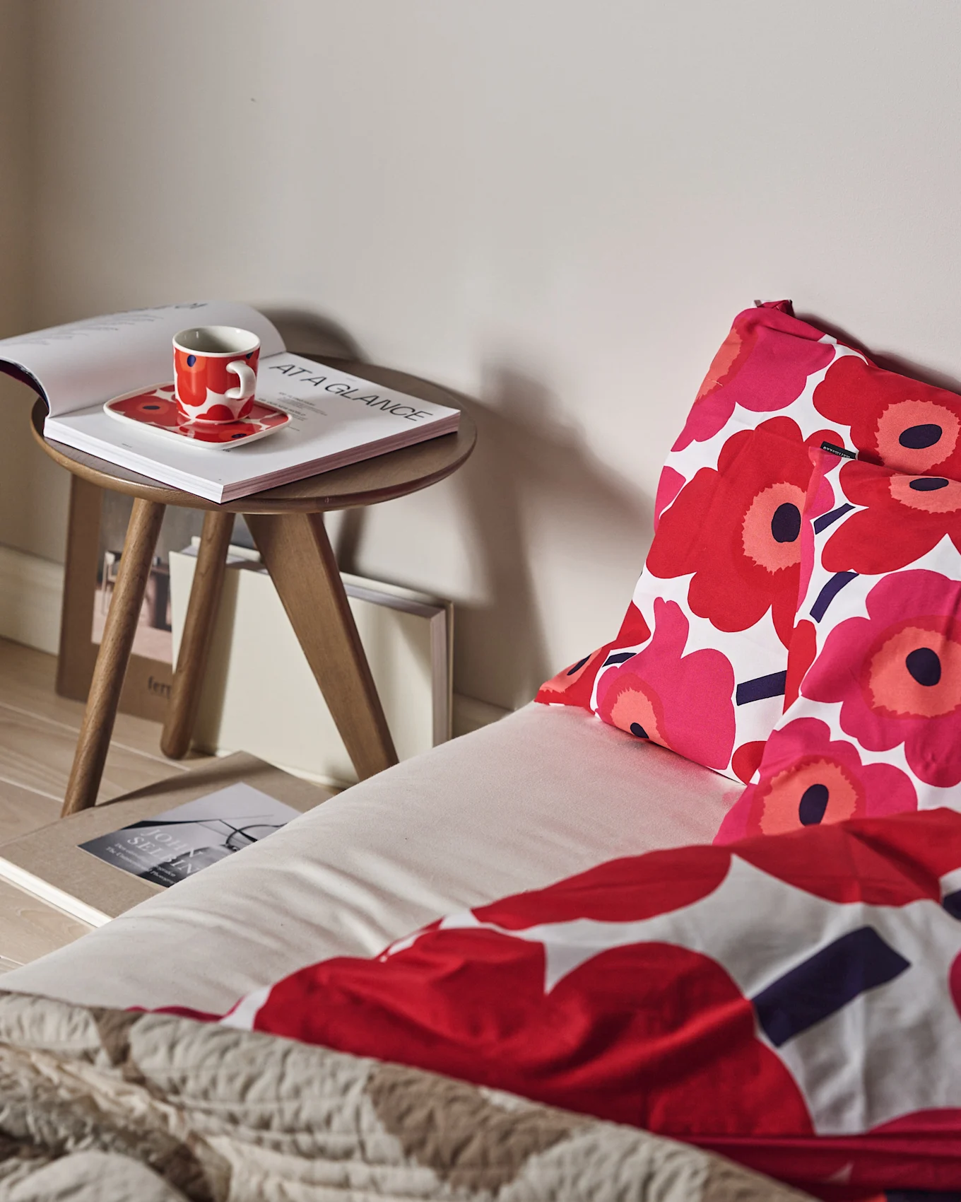Un lit avec une parure de lit florale rouge et rose et un jeté beige texturé. Une tasse rouge et un livre ouvert sur une table d'appoint en bois.