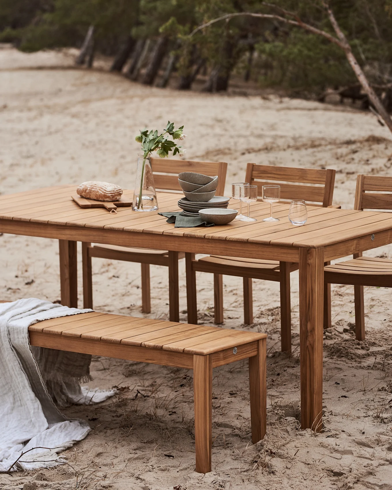 Ensemble repas d'extérieur en bois avec table et banc sur une plage de sable. La table est dressée avec de la vaisselle, des verres, du pain et un vase de fleurs.