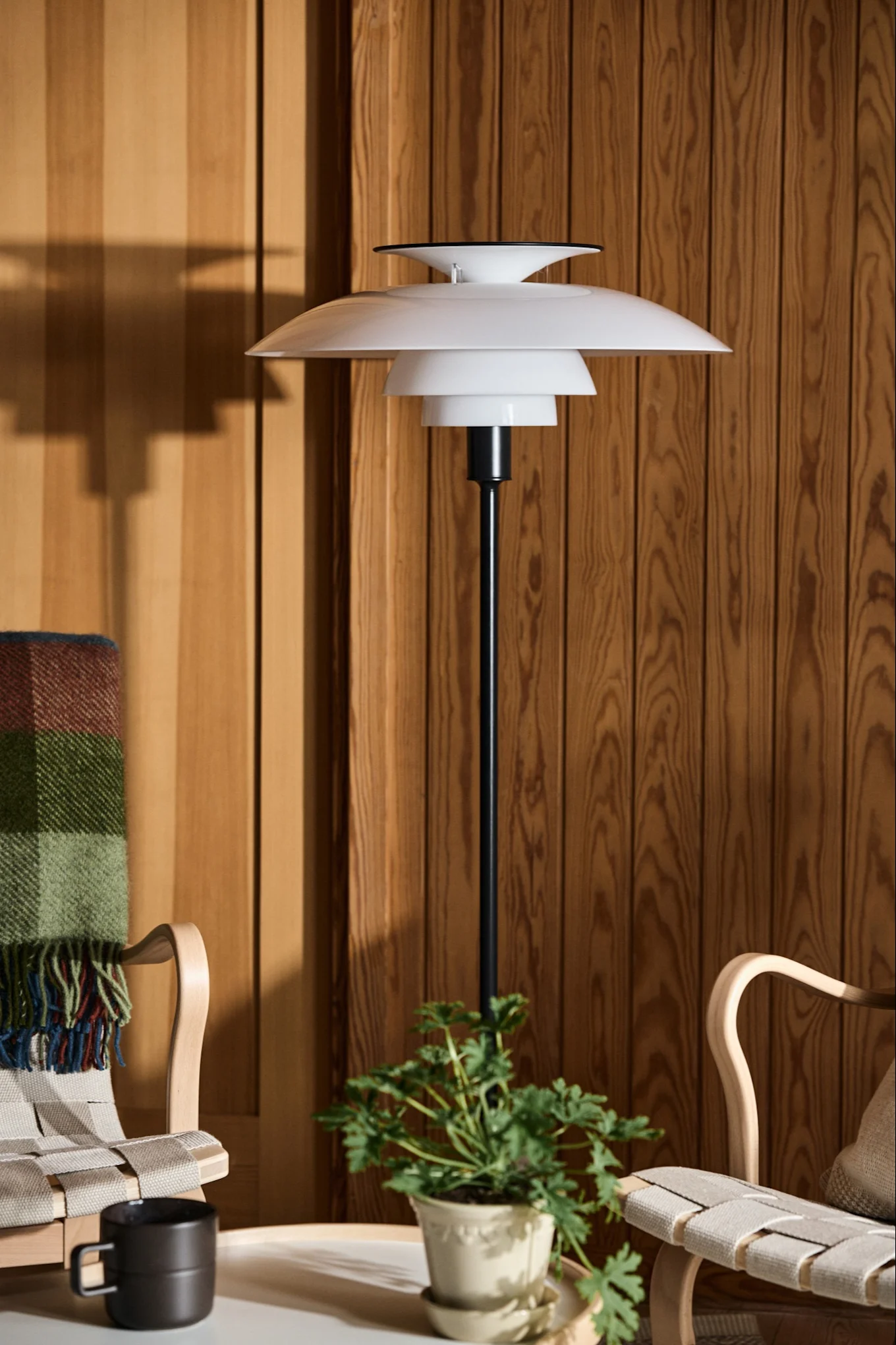 Lampadaire blanc à plusieurs niveaux à côté de deux chaises en bois et d'une plante devant un mur lambrissé.
