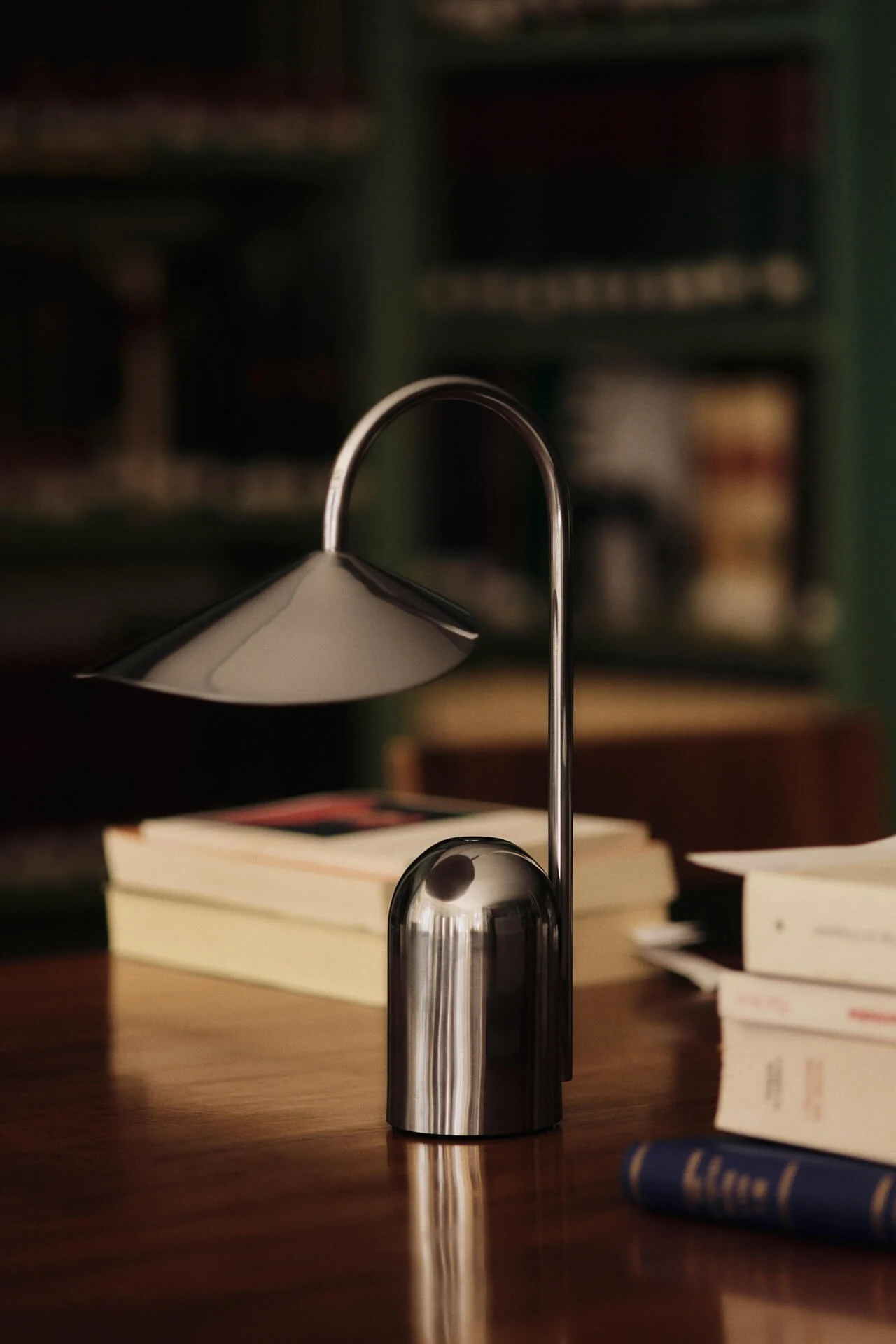 Une lampe de table argentée moderne avec une tige incurvée et un abat-jour conique sur un bureau en bois avec des livres flous en arrière-plan.