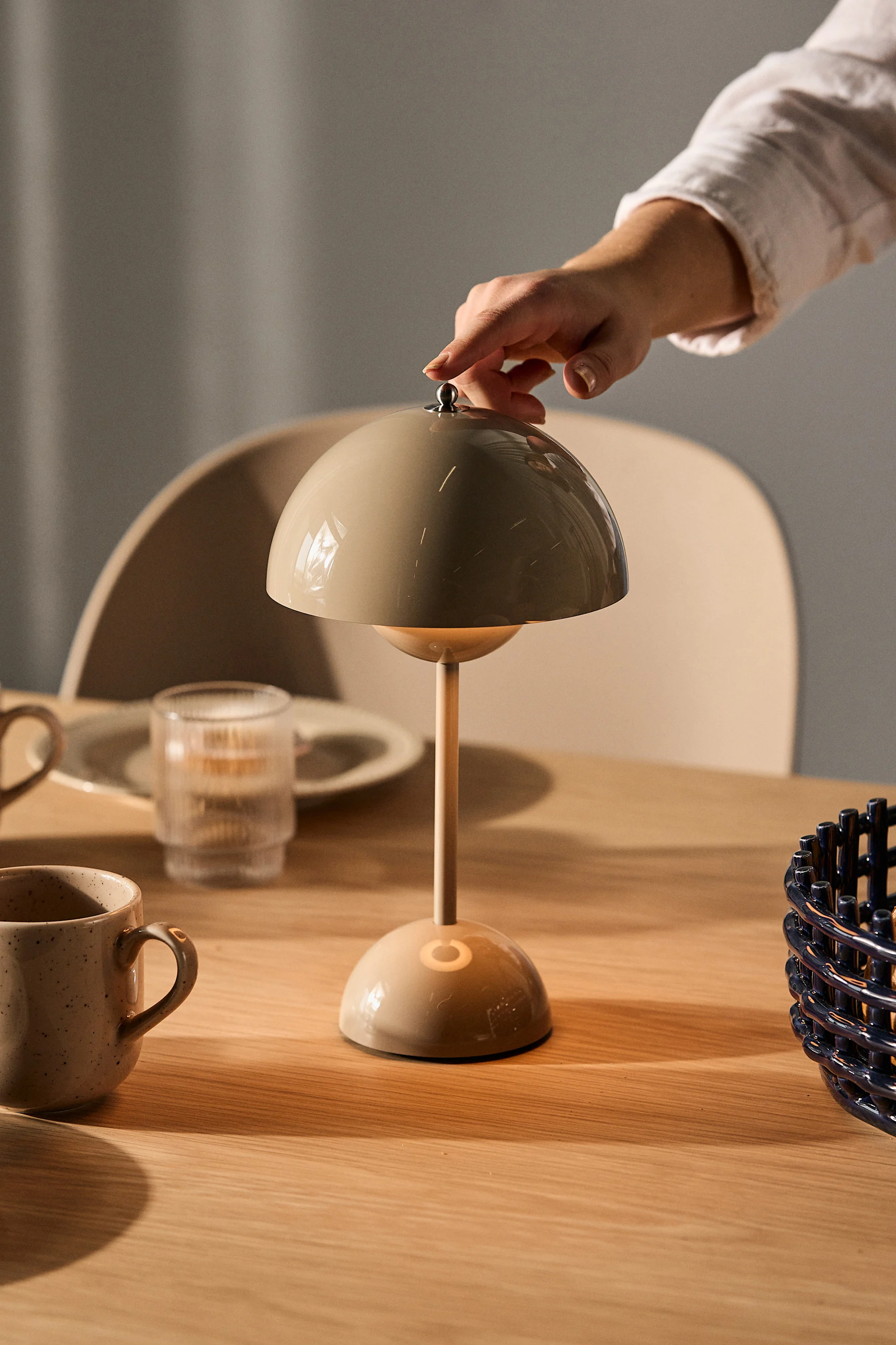 Une main ajuste une lampe de table beige en forme de champignon sur une table en bois avec des tasses et un verre.