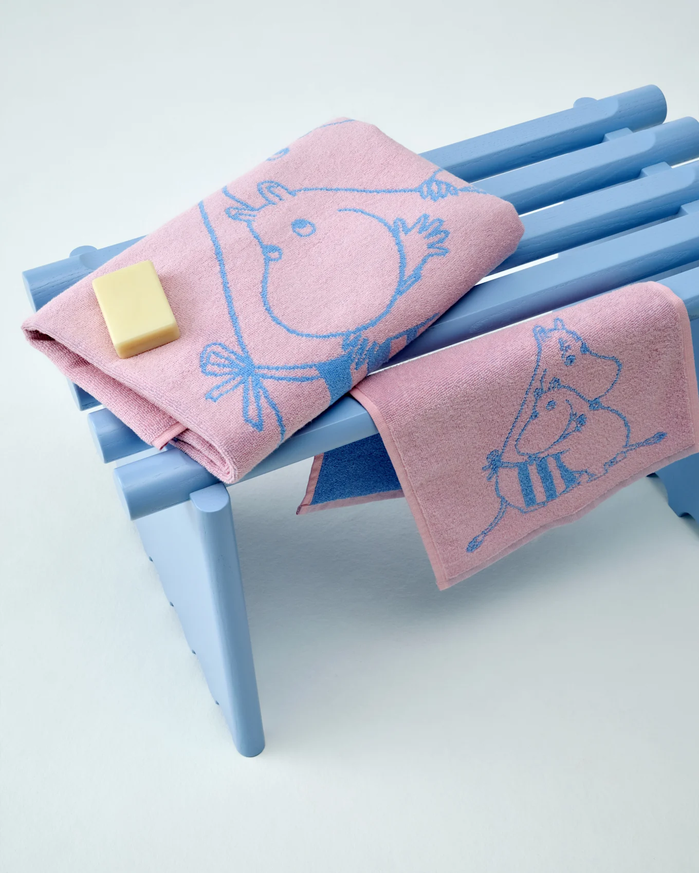 Deux serviettes Moomin roses avec motifs bleus, l'une pliée avec un pain de savon, sur un banc bleu clair.