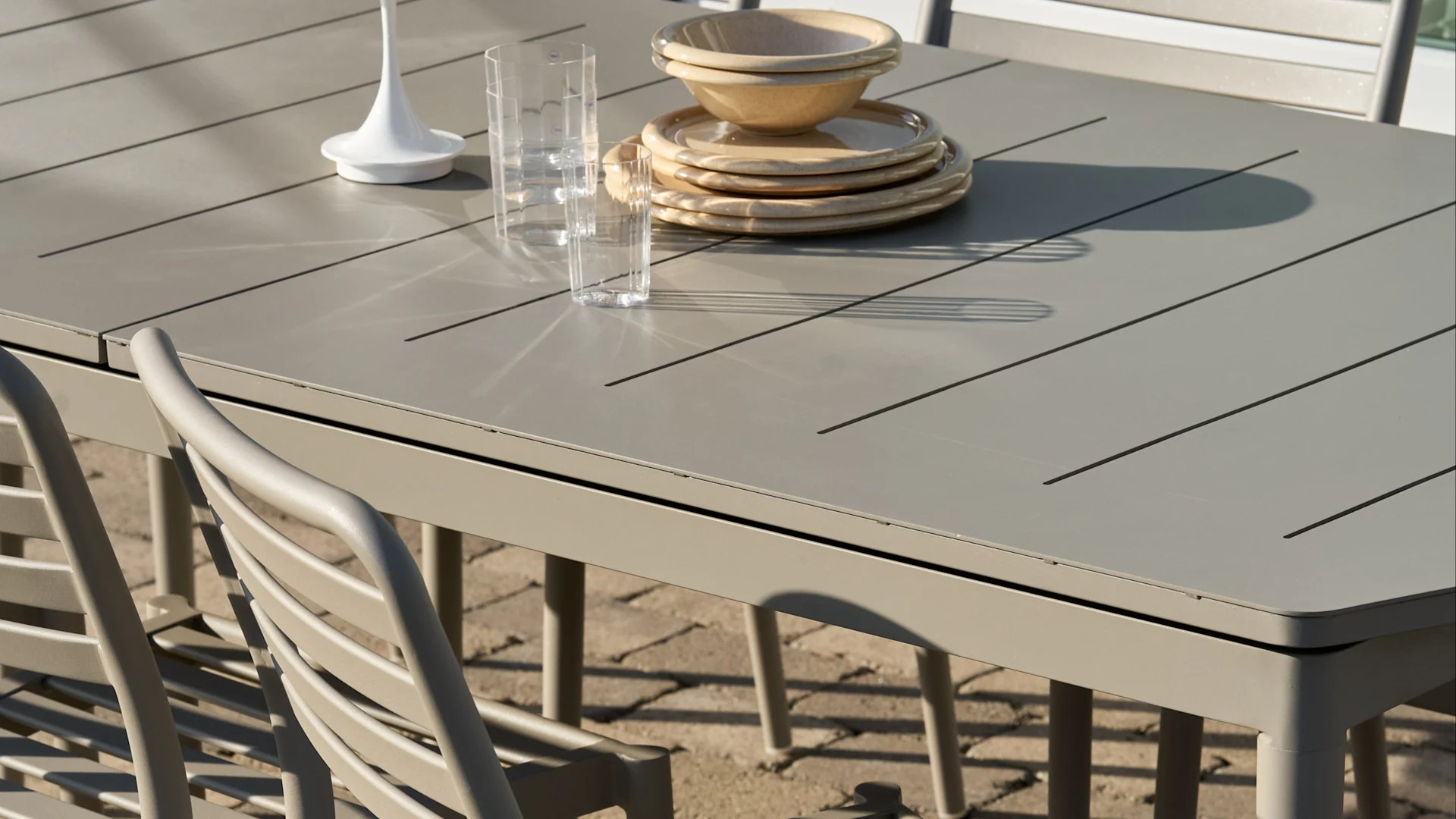 Table à manger d'extérieur à lattes gris clair avec assiettes et bols beiges empilés, deux verres transparents et un objet décoratif blanc.