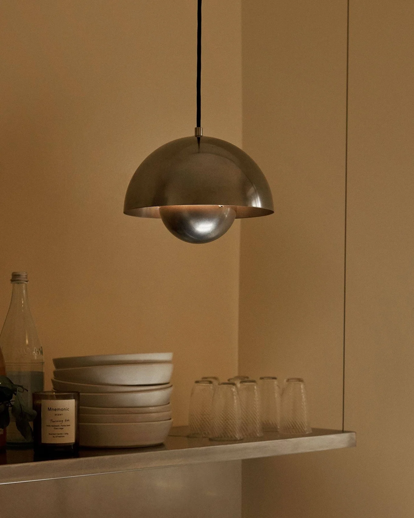 Une suspension métallique en forme de dôme éclaire une étagère avec bols, verres, bouteilles, un fruit et une bougie.