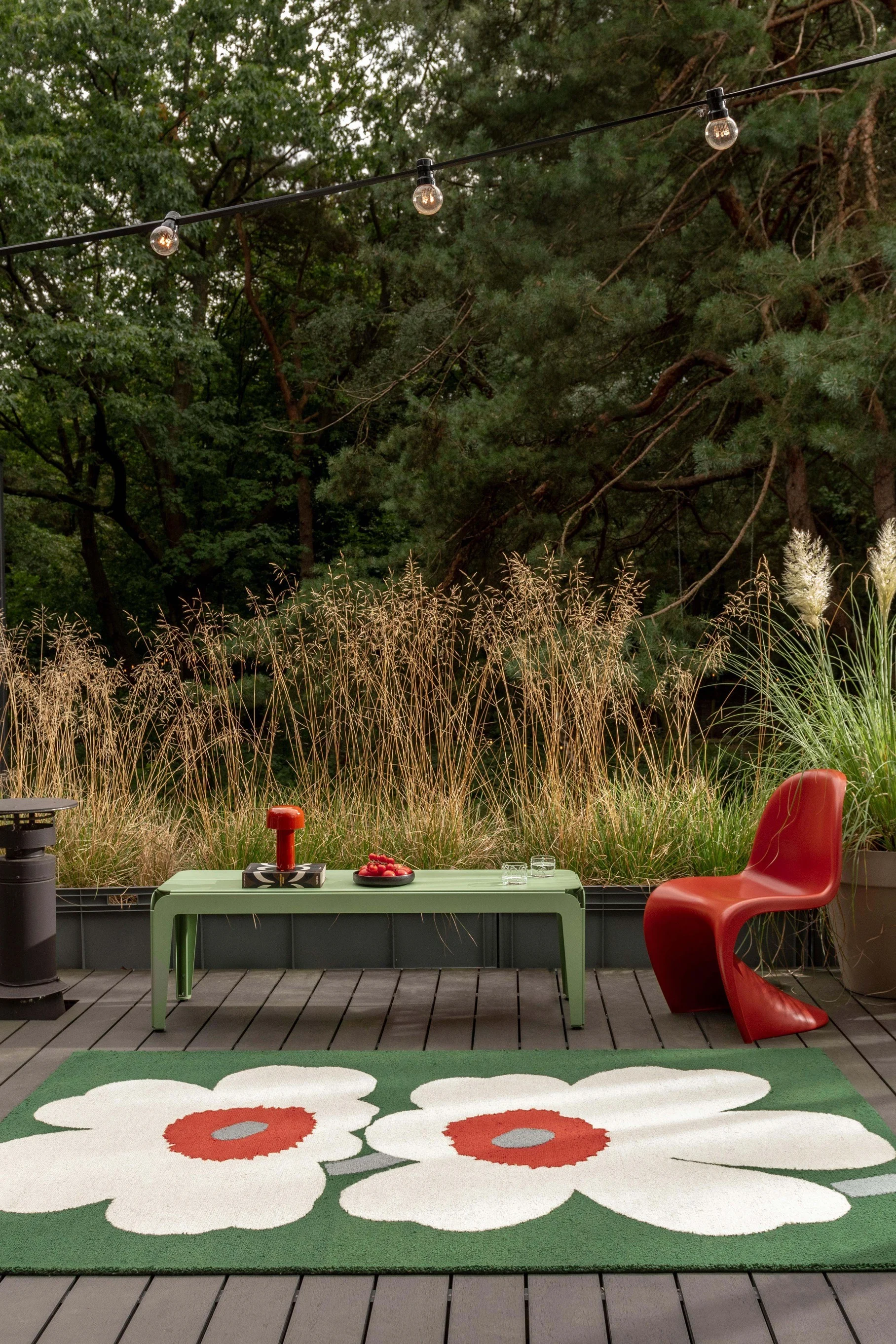 Un patio extérieur avec un tapis floral vert, une table vert clair, une chaise rouge et des guirlandes lumineuses, entouré de hautes herbes et d'arbres.