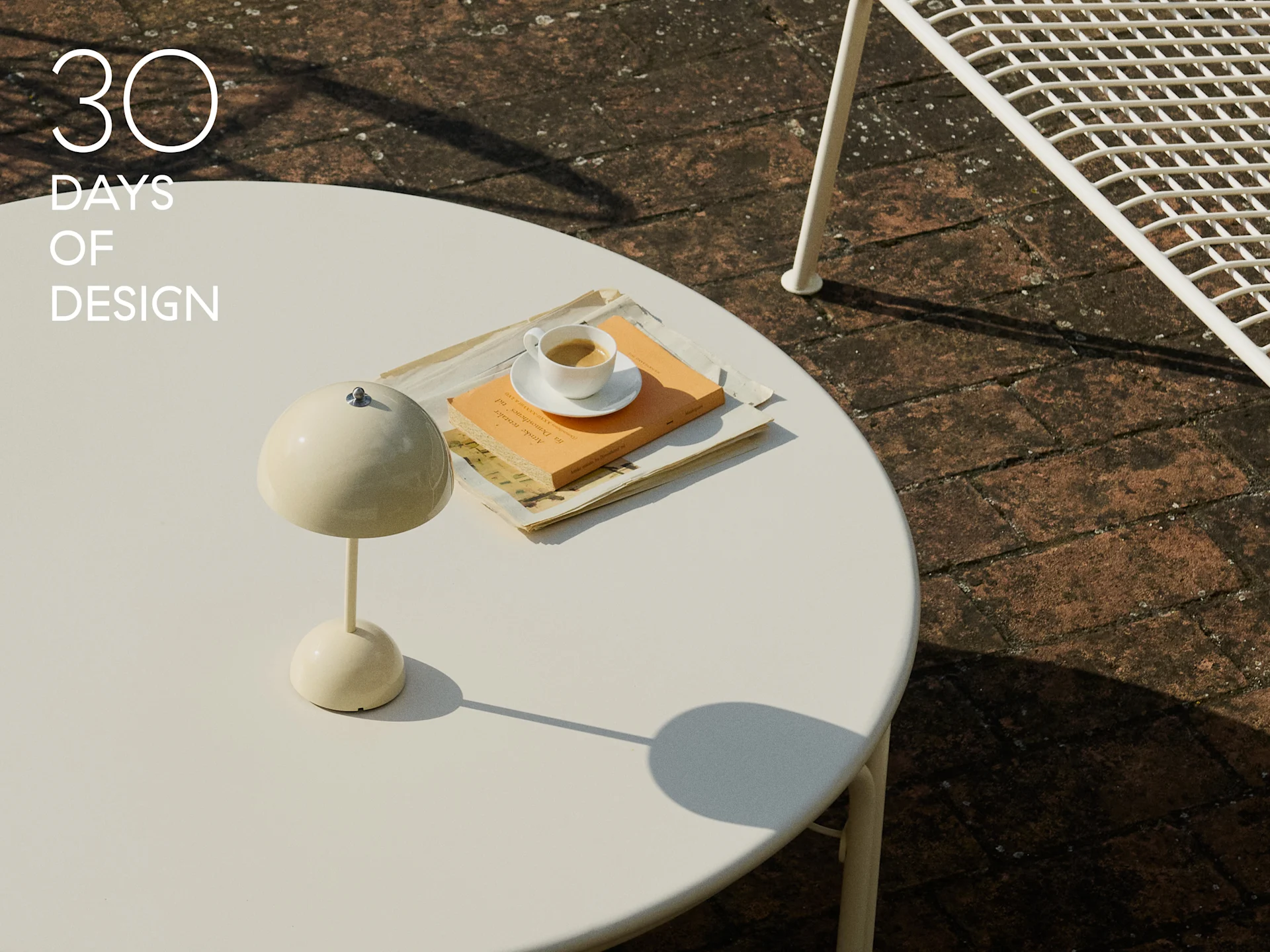 Lampe champignon crème, tasse de café et livre sur une table ronde blanche d'extérieur.