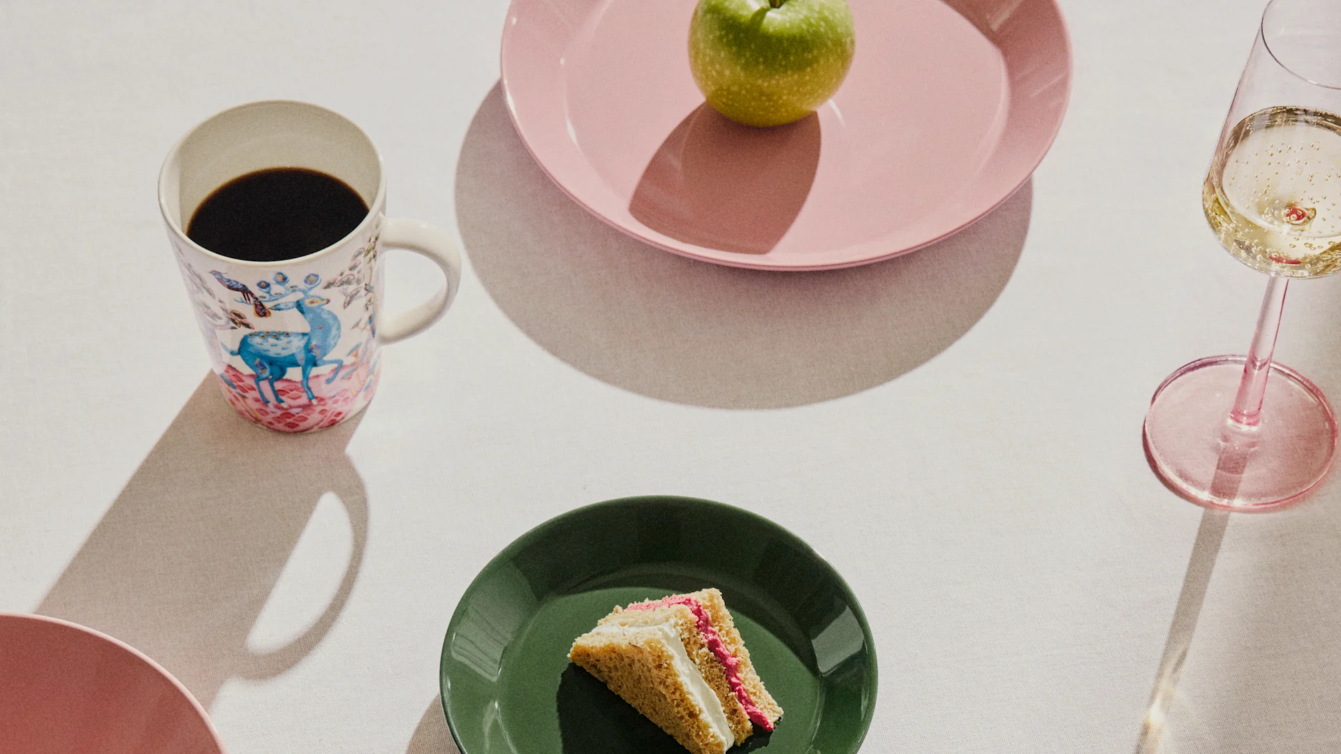 Table dressée avec une tasse à café à motifs, une pomme verte sur une assiette rose, une part de gâteau sur une assiette verte et une coupe de champagne.