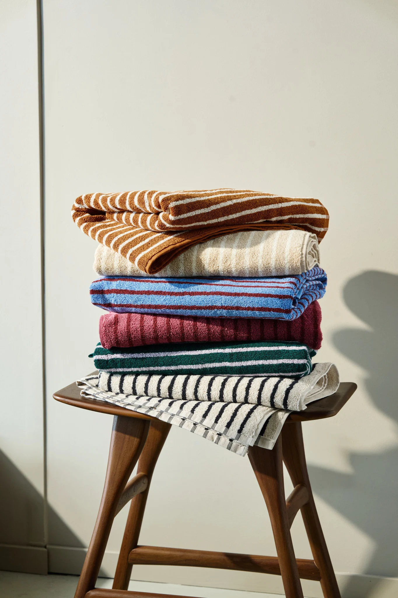 Une pile de serviettes colorées, à rayures ou unies, dans les tons marron, beige, bleu, bordeaux, vert et noir, posée sur un tabouret en bois.