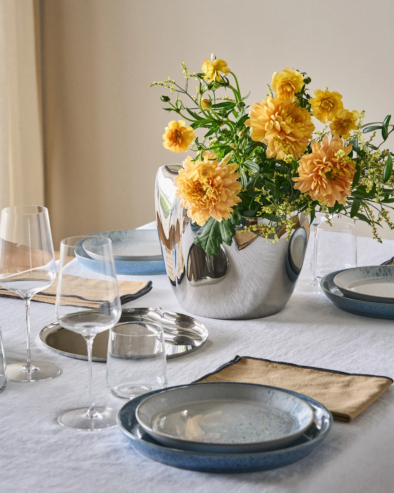 Une table à manger dressée avec des assiettes bleues, des serviettes beiges, des verres transparents et un vase argenté avec des fleurs orange-jaunes sur une nappe blanche.