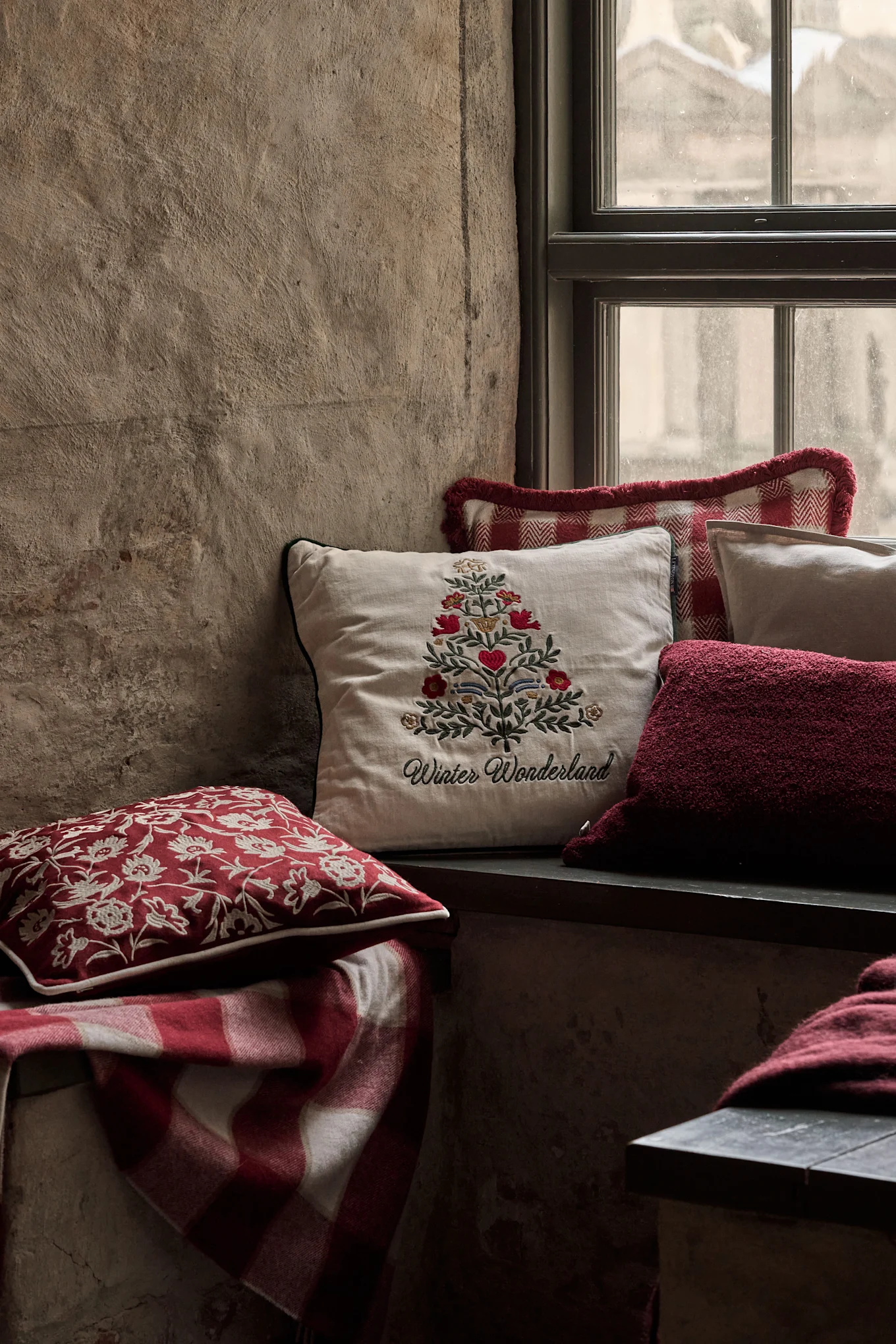Coin de fenêtre douillet avec des coussins festifs, dont un avec un sapin de Noël brodé et 'Winter Wonderland', et une couverture à carreaux rouge.