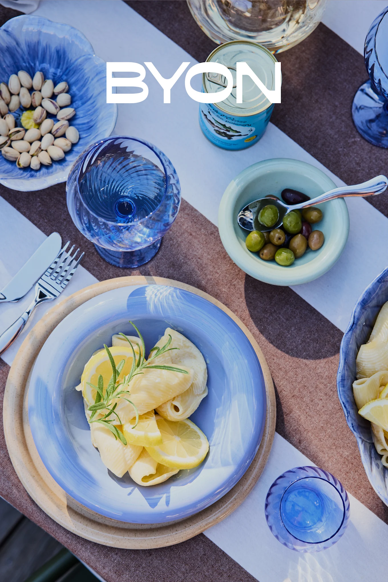 Vue plongeante d'une table à manger avec de la vaisselle bleue, comprenant une assiette de pâtes au citron et au romarin, des bols de pistaches et d'olives, et des verres bleus. Le logo BYON est superposé.