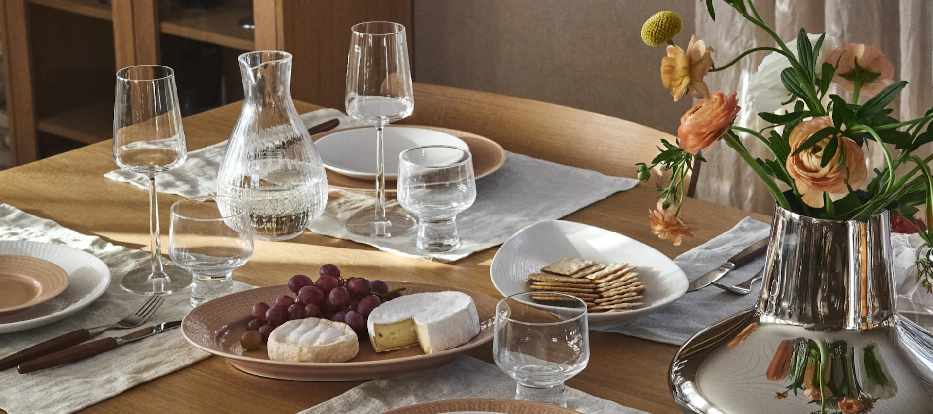 Une élégante table à manger dressée avec des verres transparents, une carafe d'eau, des assiettes rose pâle et blanches, des couverts, un plateau de fromages et de raisins, des crackers et un vase argenté avec des fleurs couleur pêche.