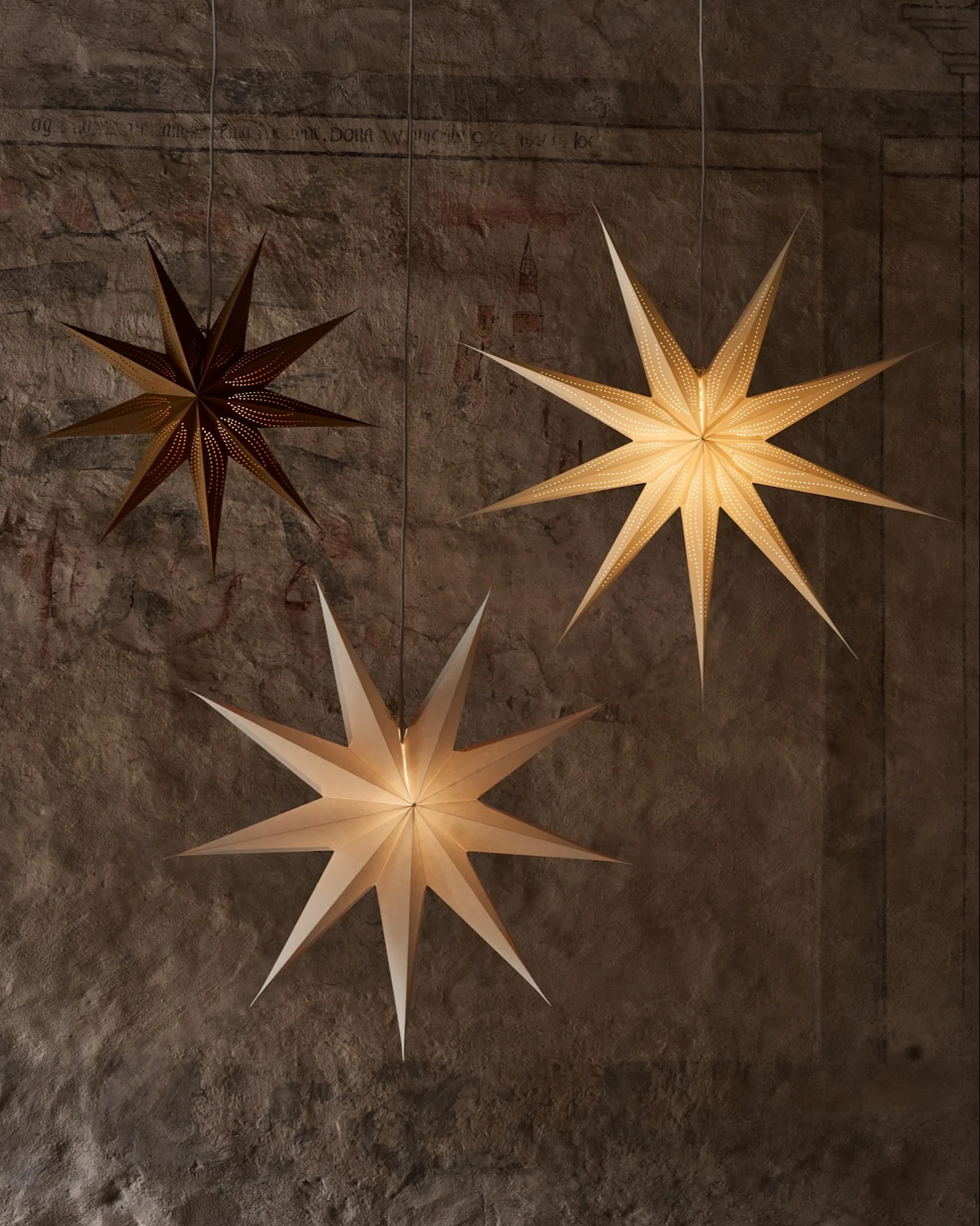 Trois lanternes en papier en forme d'étoile sont suspendues devant un mur texturé. Une lanterne sombre est perforée, deux plus claires éclairent la scène.