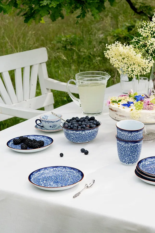 Table de jardin avec vaisselle bleue et blanche, myrtilles et mûres fraîches, carafe de limonade et gâteau décoré de fleurs.