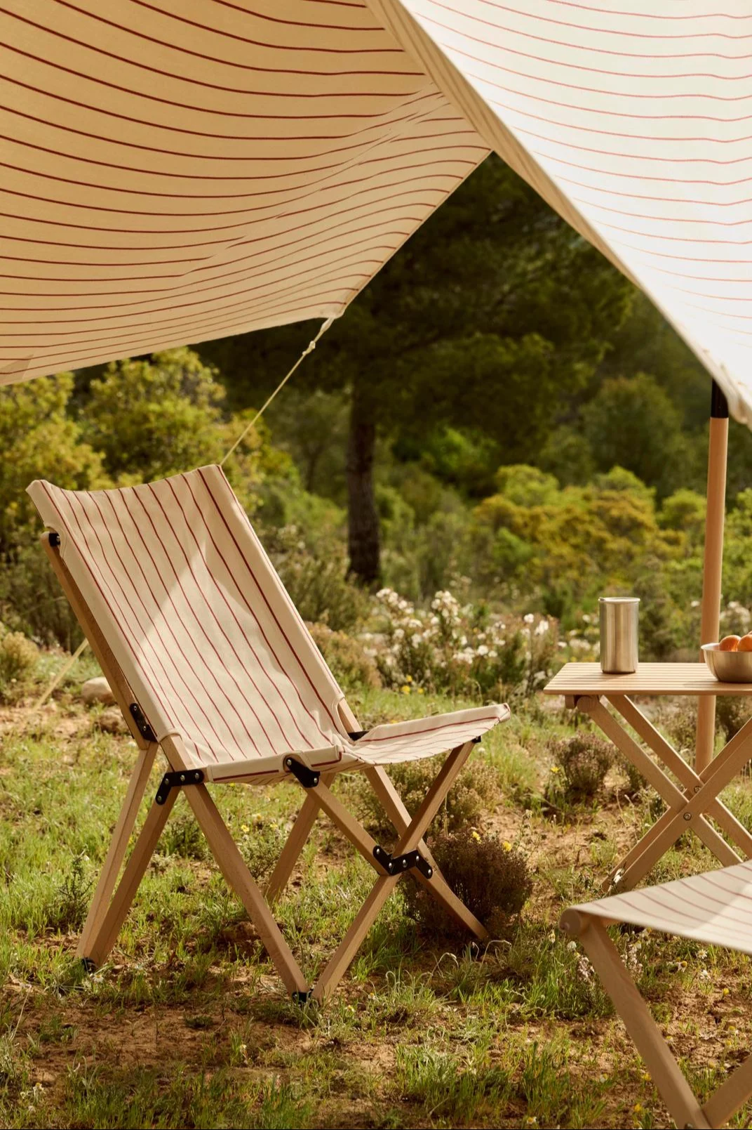 Un store rayé ombrage une chaise pliante en bois rayée et une table sur laquelle sont posés un thermos et des oranges, à l'extérieur.