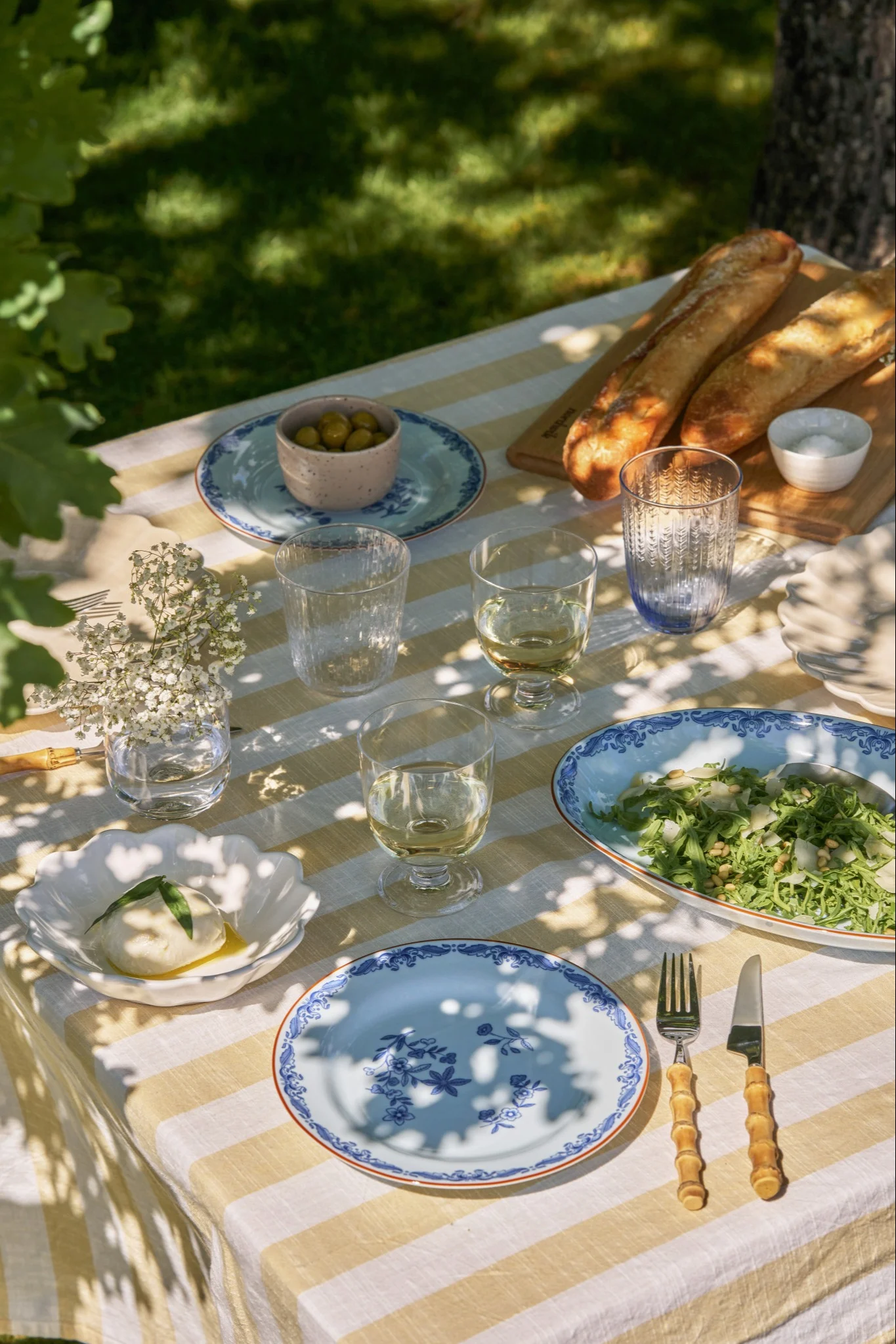 Table de pique-nique extérieure dressée avec nappe rayée jaune et blanche, burrata, salade, baguettes, olives et assiettes bleues.