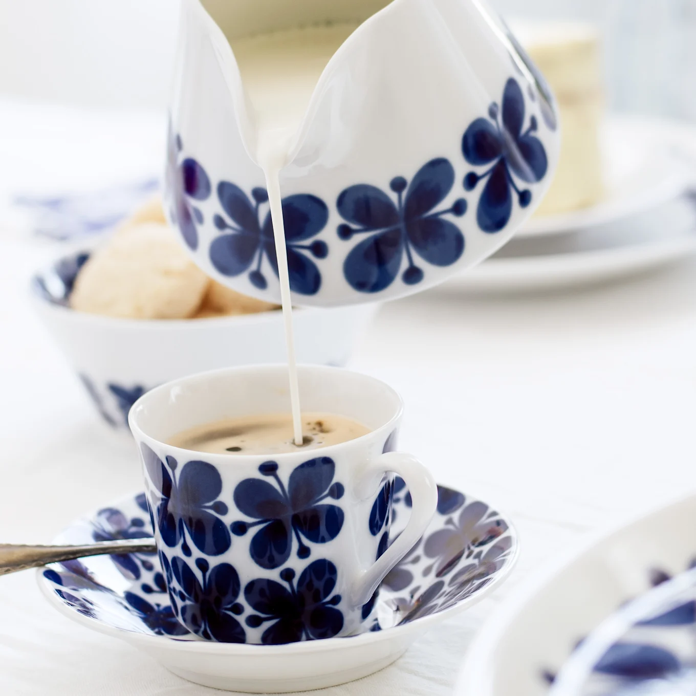 Du lait est versé d'un crémier floral bleu et blanc dans une tasse à café assortie sur une soucoupe, avec des biscuits en arrière-plan.