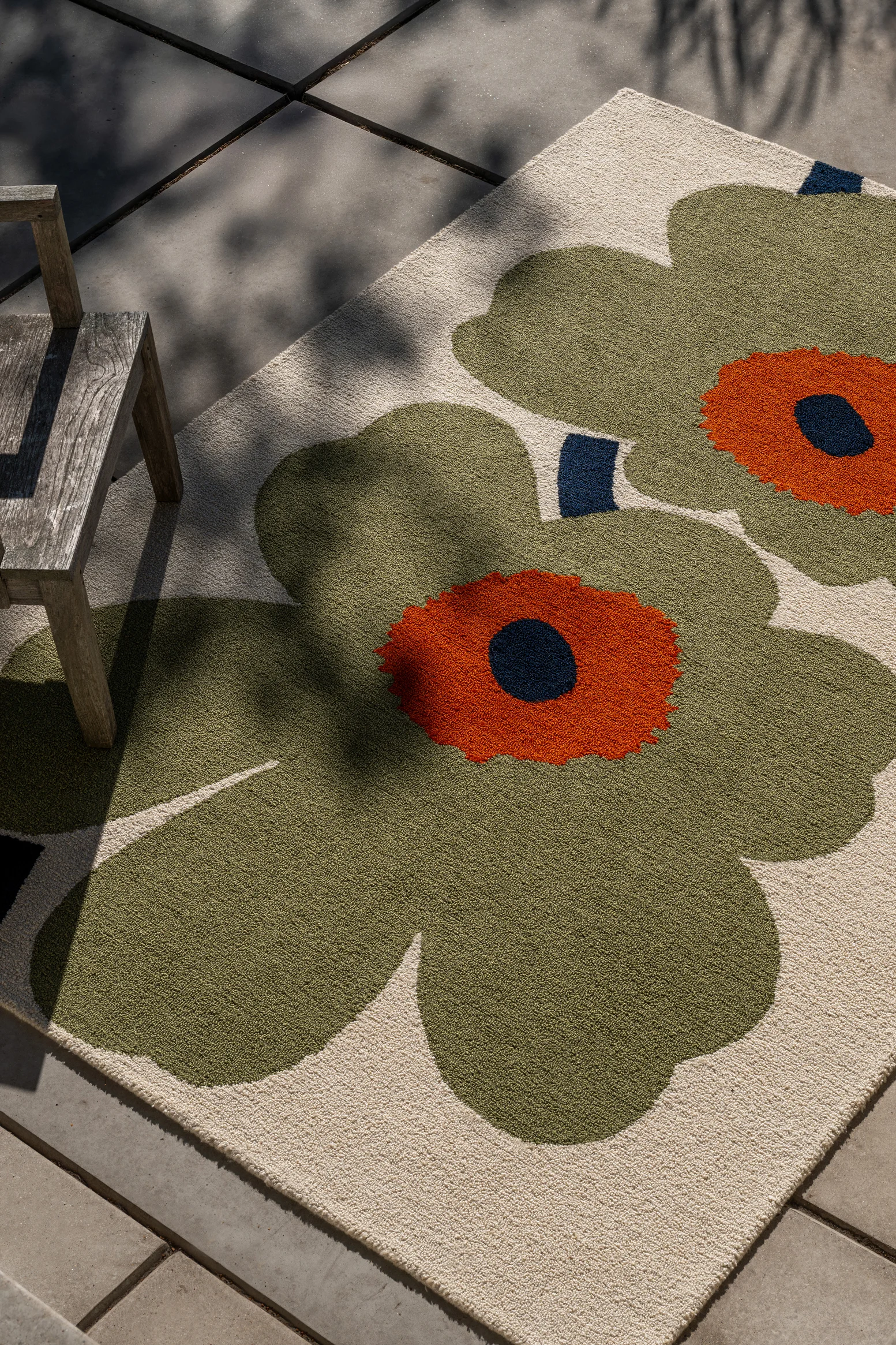 Tapis à motifs floraux verts et oranges sur fond clair, sous une chaise en bois sur patio, avec ombres de feuilles.