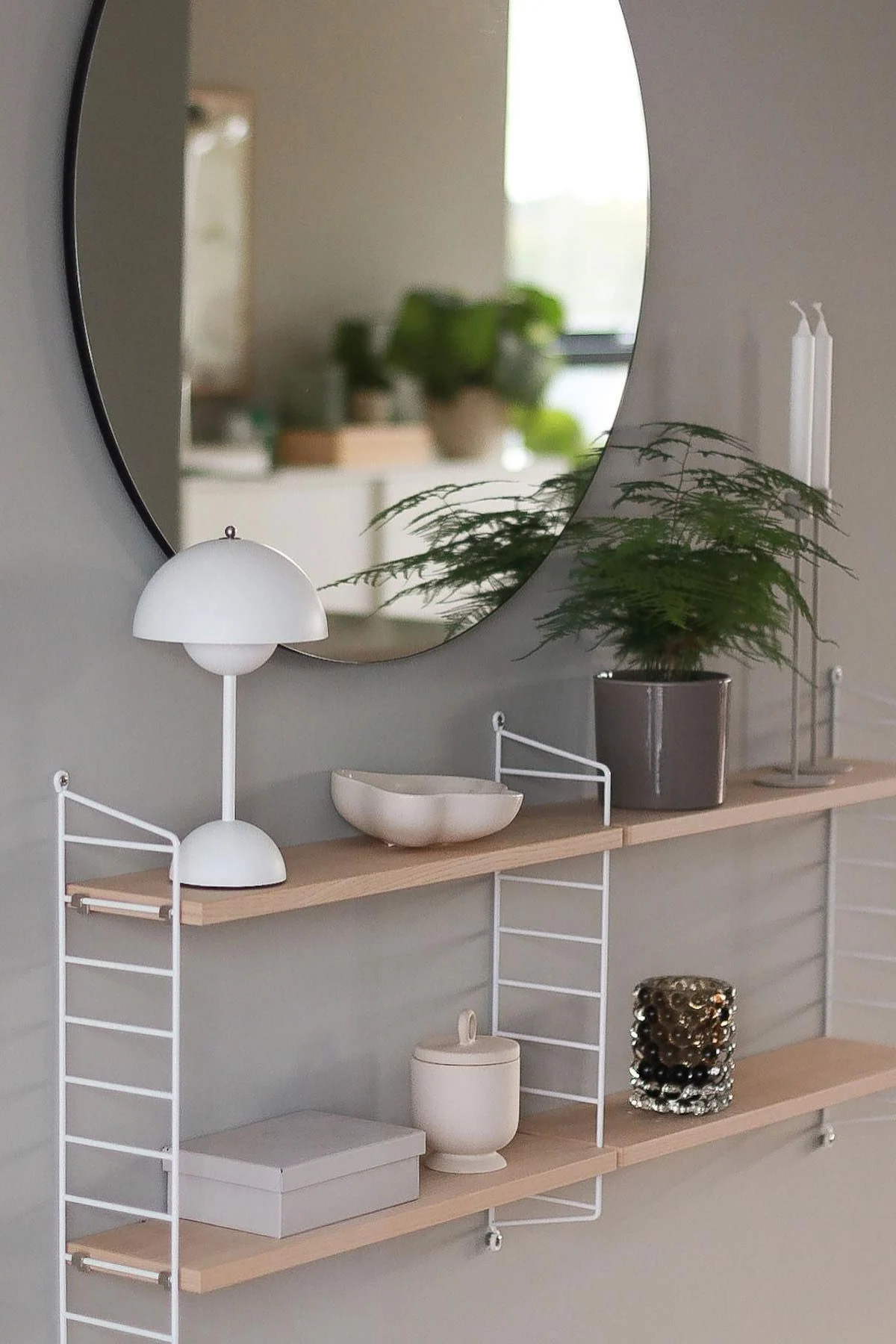 Un miroir rond au-dessus d'étagères murales blanches, avec une lampe champignon, une fougère, des objets décoratifs et des bougies.
