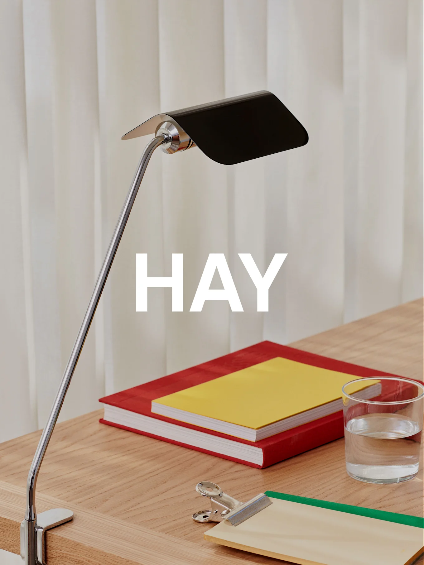 Une lampe de bureau chromée avec un abat-jour noir illumine un bureau en bois avec deux livres, un verre d'eau et un presse-papiers.