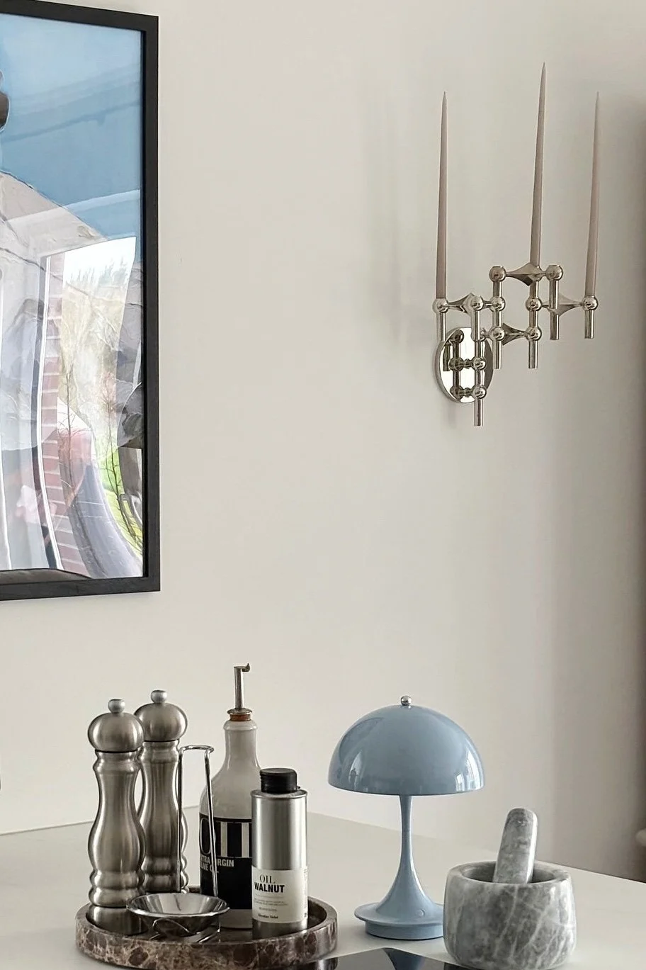 Une applique murale argentée à trois bougies est suspendue au-dessus d'une table blanche sur laquelle sont disposés divers ustensiles de cuisine : des moulins, des bouteilles d'huile, une lampe en forme de champignon bleu, ainsi qu'un mortier et un pilon en marbre.