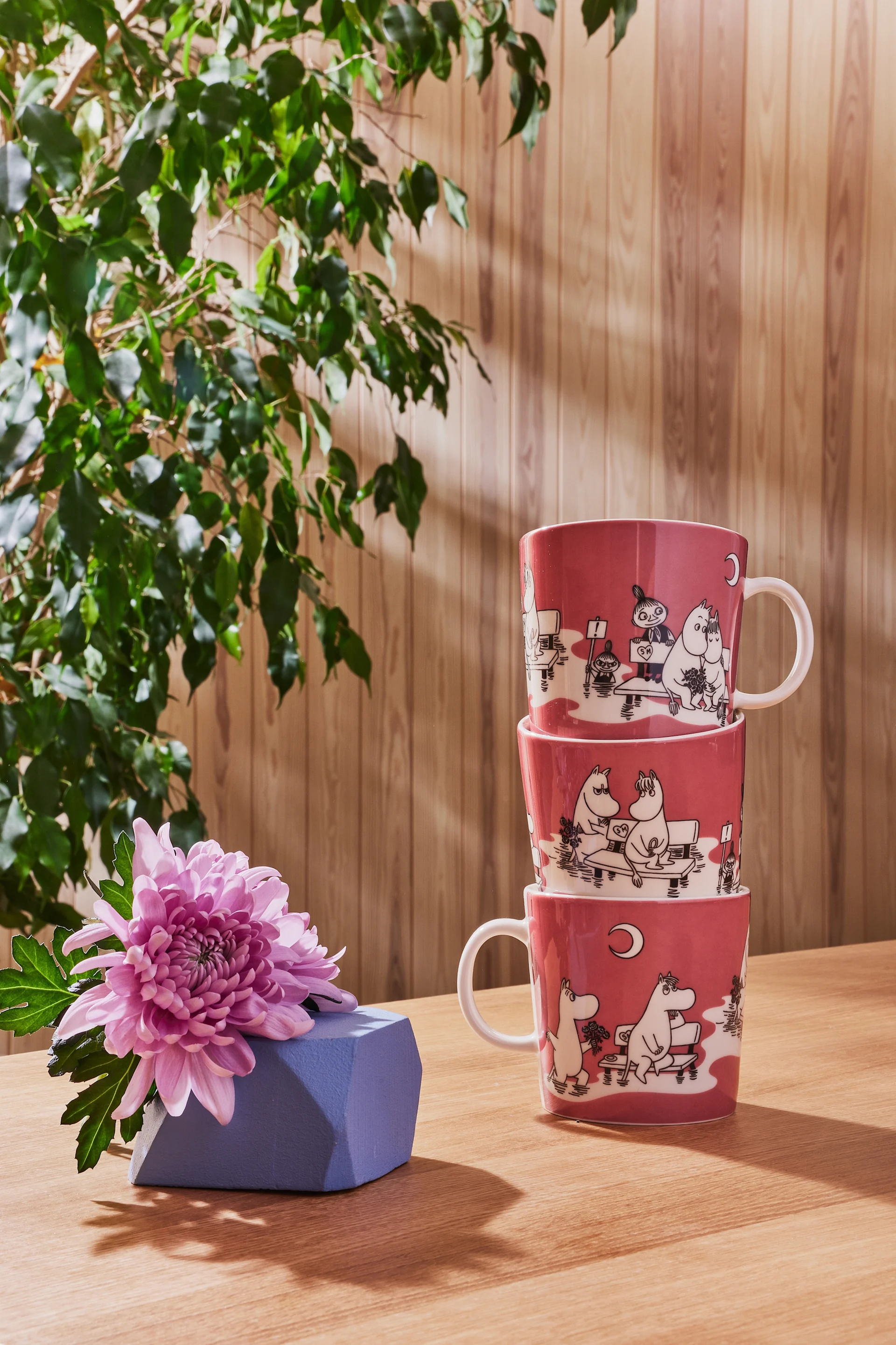 Trois tasses Moomin roses de la collection couleur sont empilées sur une table, avec un cadeau posé à côté.