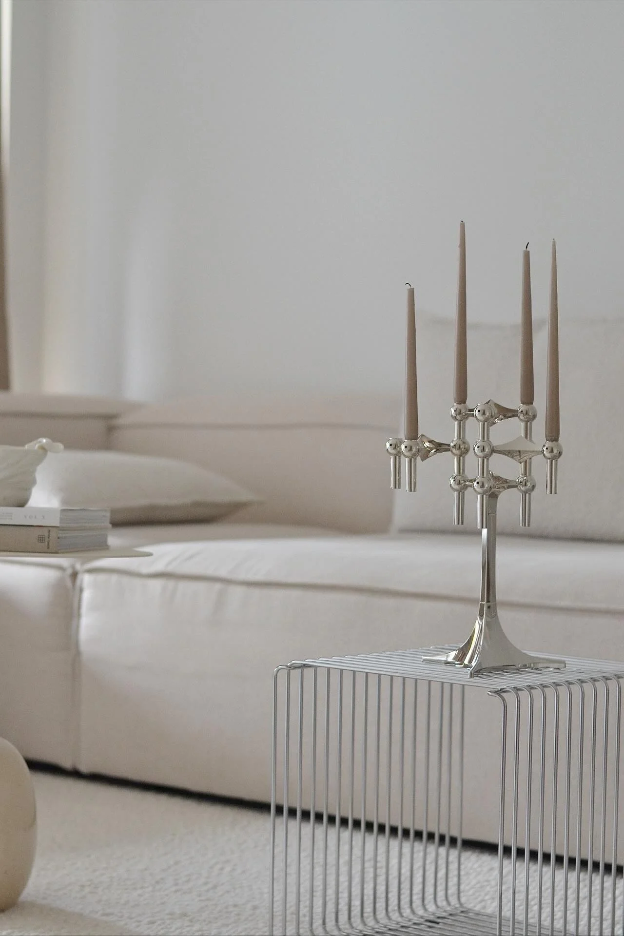 Un candélabre en argent orné de cinq bougies brun clair trône sur une table d'appoint minimaliste en métal. Un canapé de couleur claire se trouve à l'arrière-plan.