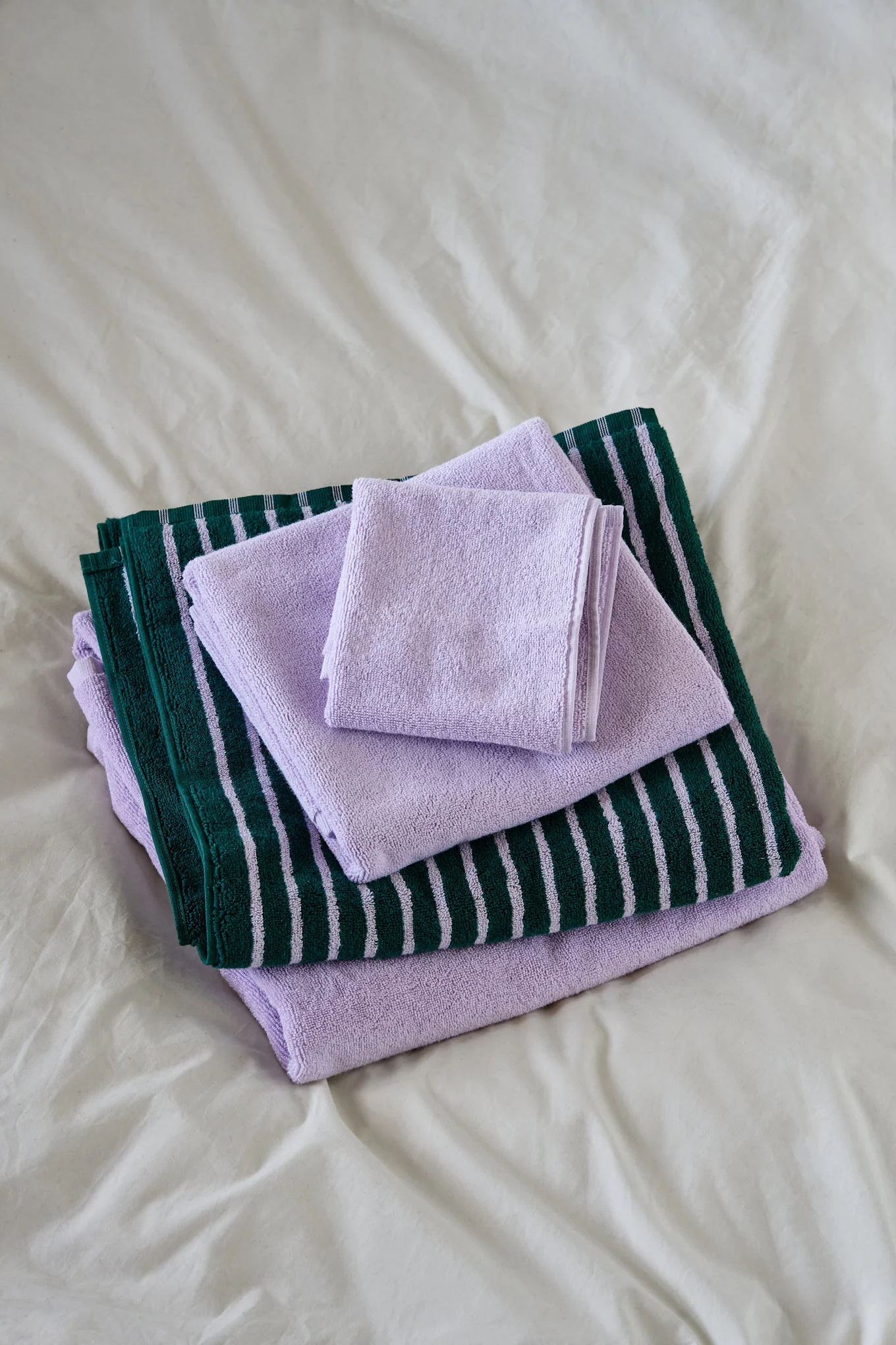 Des serviettes pliées et empilées : violet clair et vert foncé à rayures violettes, sur un drap blanc.