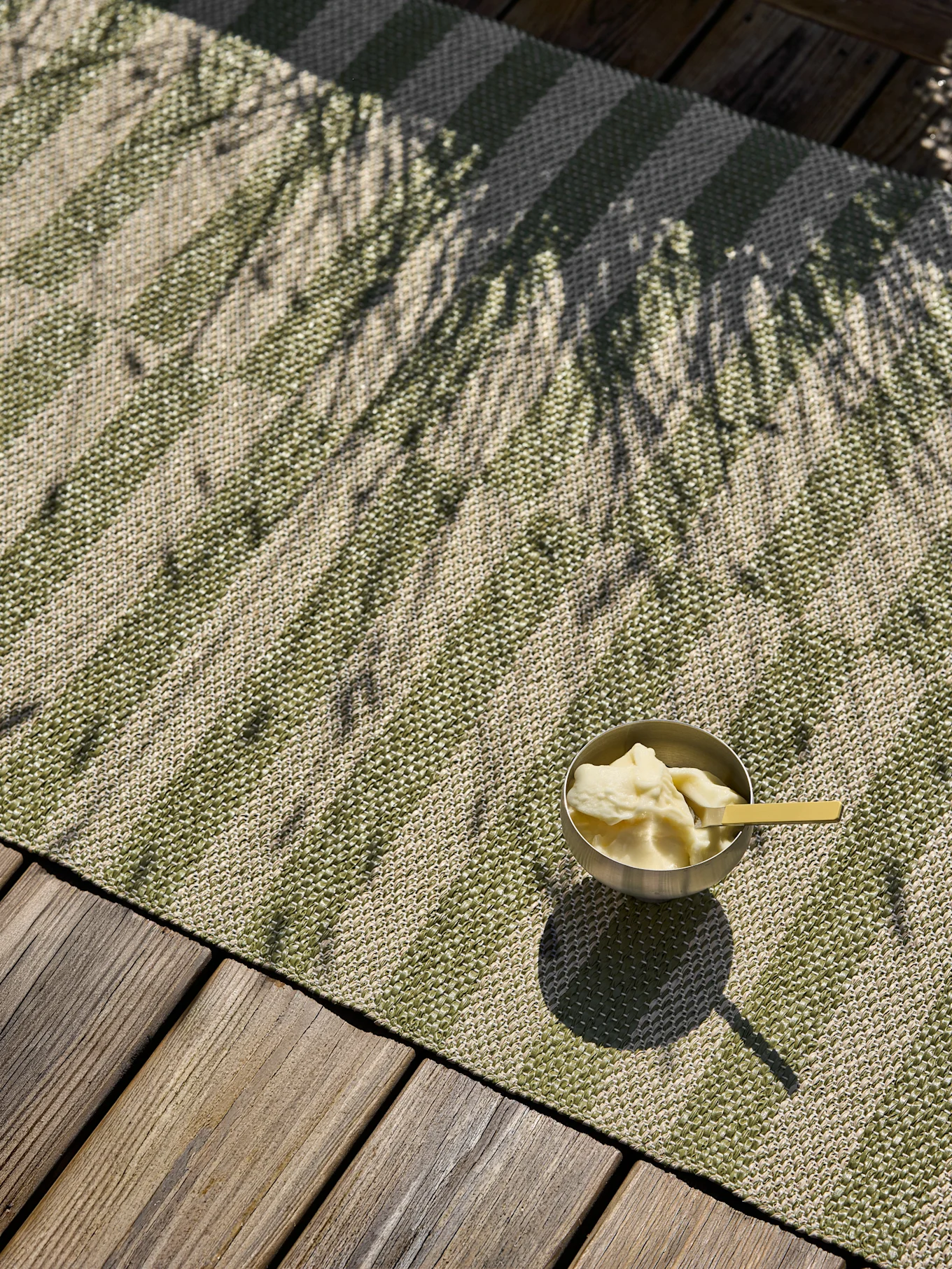 Un bol d'argent de dessert avec une cuillère jaune est posé sur un tapis extérieur rayé vert et beige sur une terrasse en bois, moucheté d'ombres de palmiers.