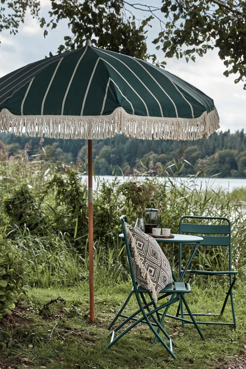 Décoration bohème pour le jardin : le parasol vert à franges de House Doctor dans un jardin, à côté d'un petit coin salon.
