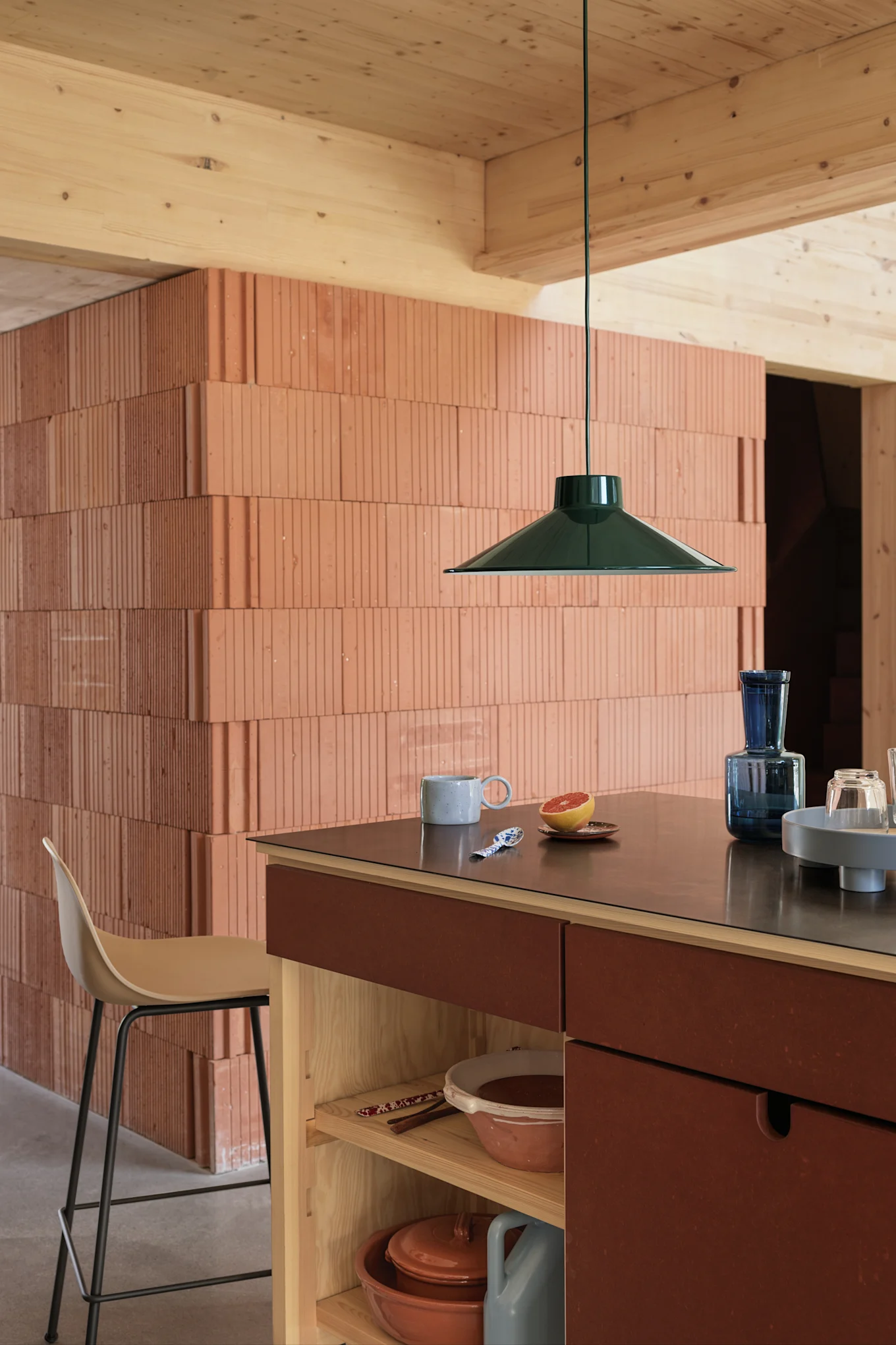 Suspension vert foncé au-dessus d'un îlot de cuisine avec un plan de travail foncé, une tasse, un pamplemousse et des verres bleus, mur en briques texturées.