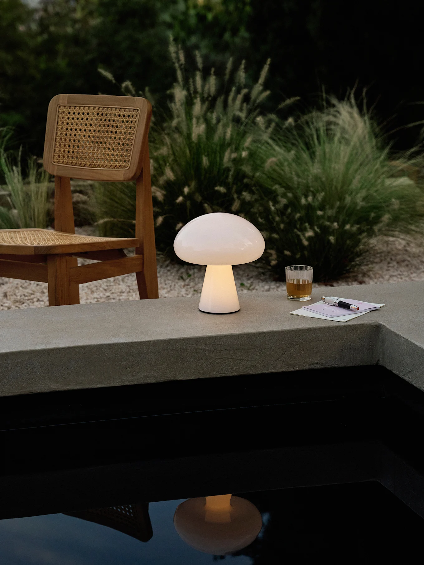 Une lampe lumineuse en forme de champignon sur un rebord en béton près d'une piscine, avec une chaise en bois, une boisson et un stylo.