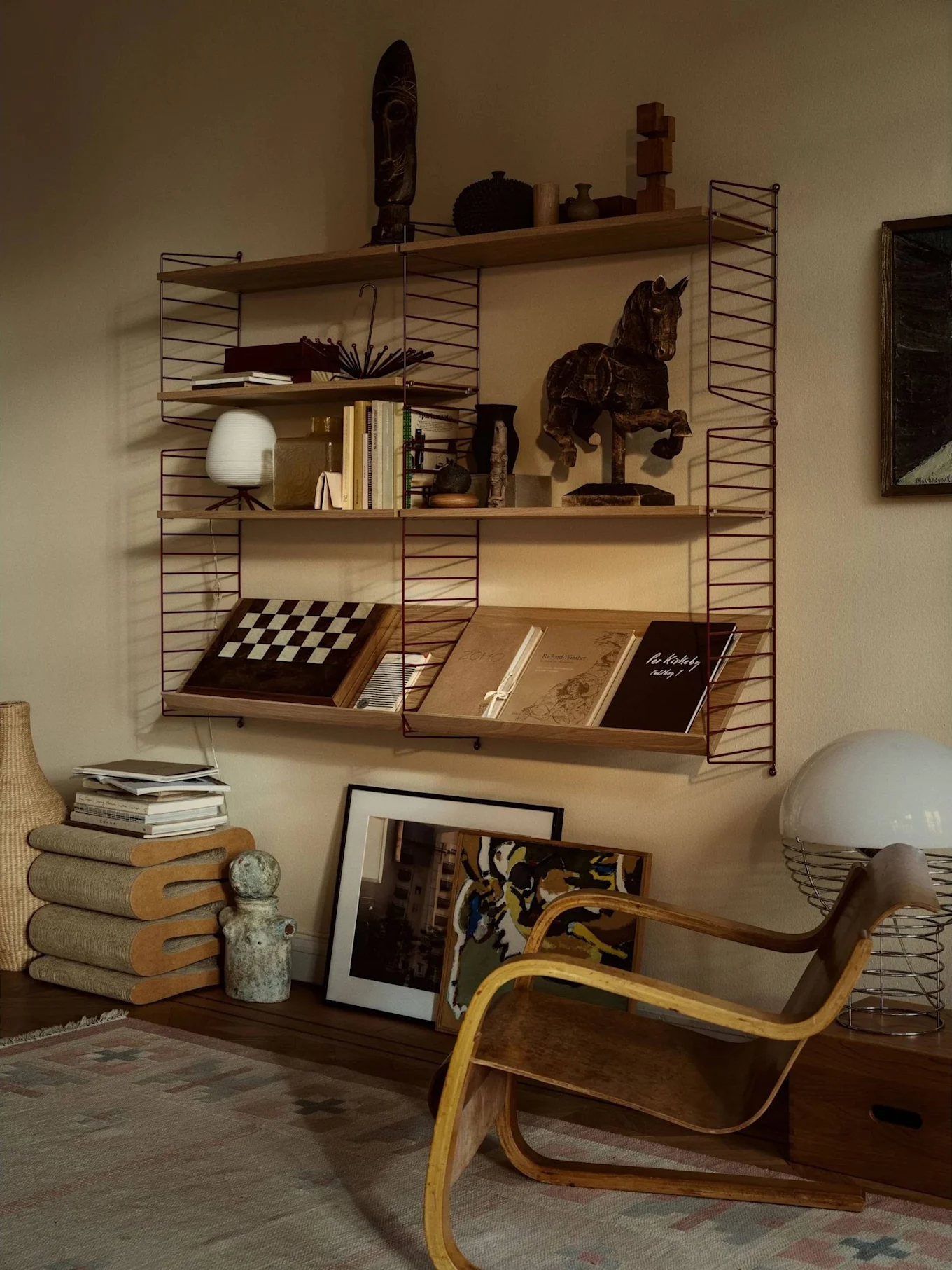 Une pièce chaleureuse avec une étagère remplie de livres, d'art et de décorations, à côté d'un fauteuil en bois courbé et d'une grande lampe blanche.