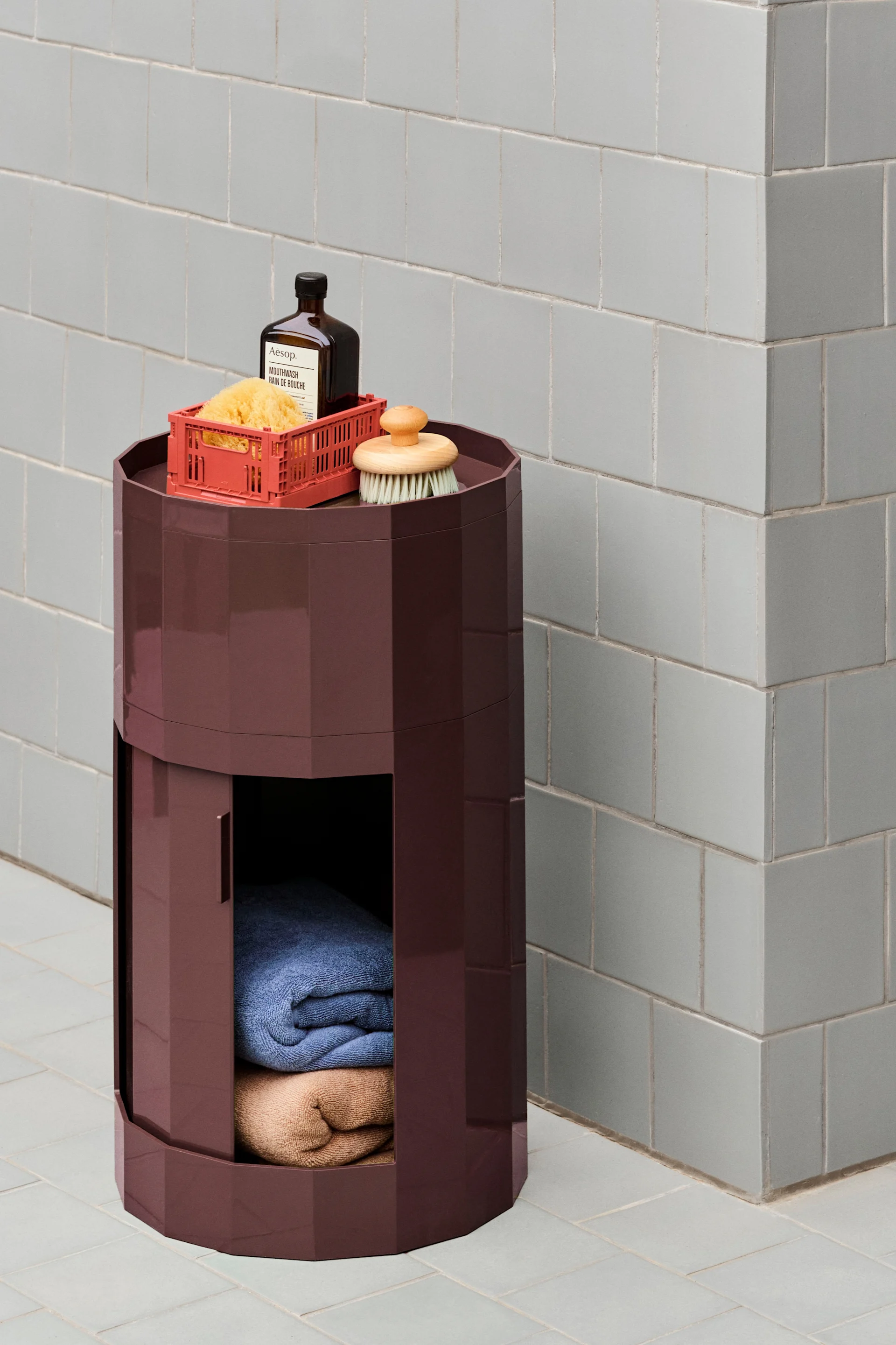 Une unité de rangement hexagonale rouge foncé avec des produits de bain sur le dessus et des serviettes pliées à l'intérieur, contre un mur carrelé gris clair.