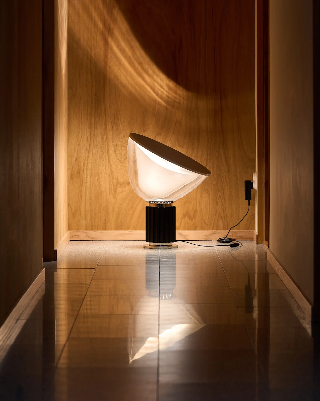 Lampe moderne avec abat-jour semi-circulaire blanc opaque et base noire striée dans couloir boisé au sol réfléchissant. Motif lumineux mural.