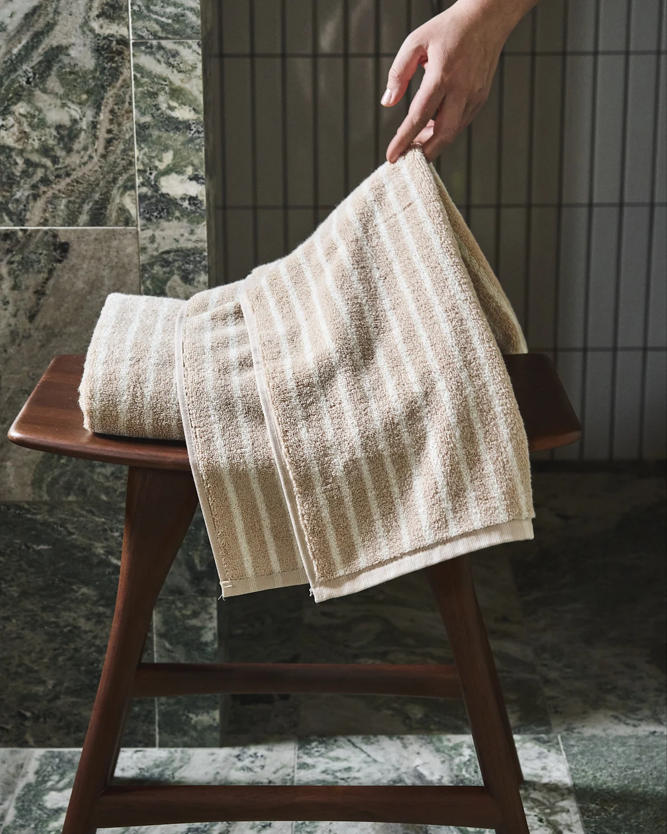 Main ajustant une serviette rayée beige et blanche sur un tabouret en bois.