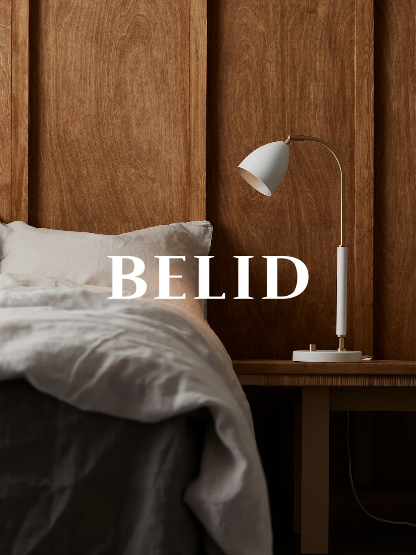 Lampe blanche et laiton sur une table de chevet en bois, avec le texte 'BELID' sur un lit aux draps de lin.