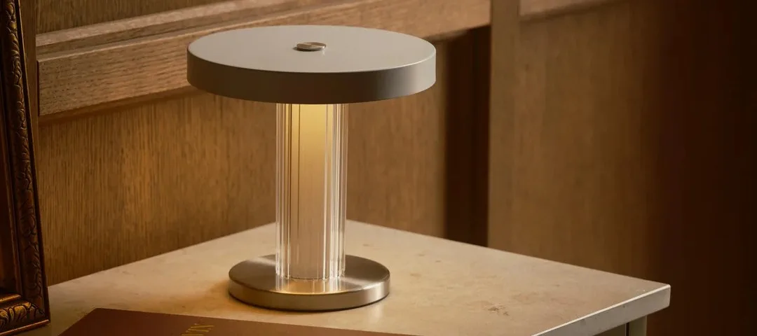 Lampe de table moderne allumée avec un corps cannelé transparent et un plateau gris, à côté de dossiers marron sur un meuble vert.