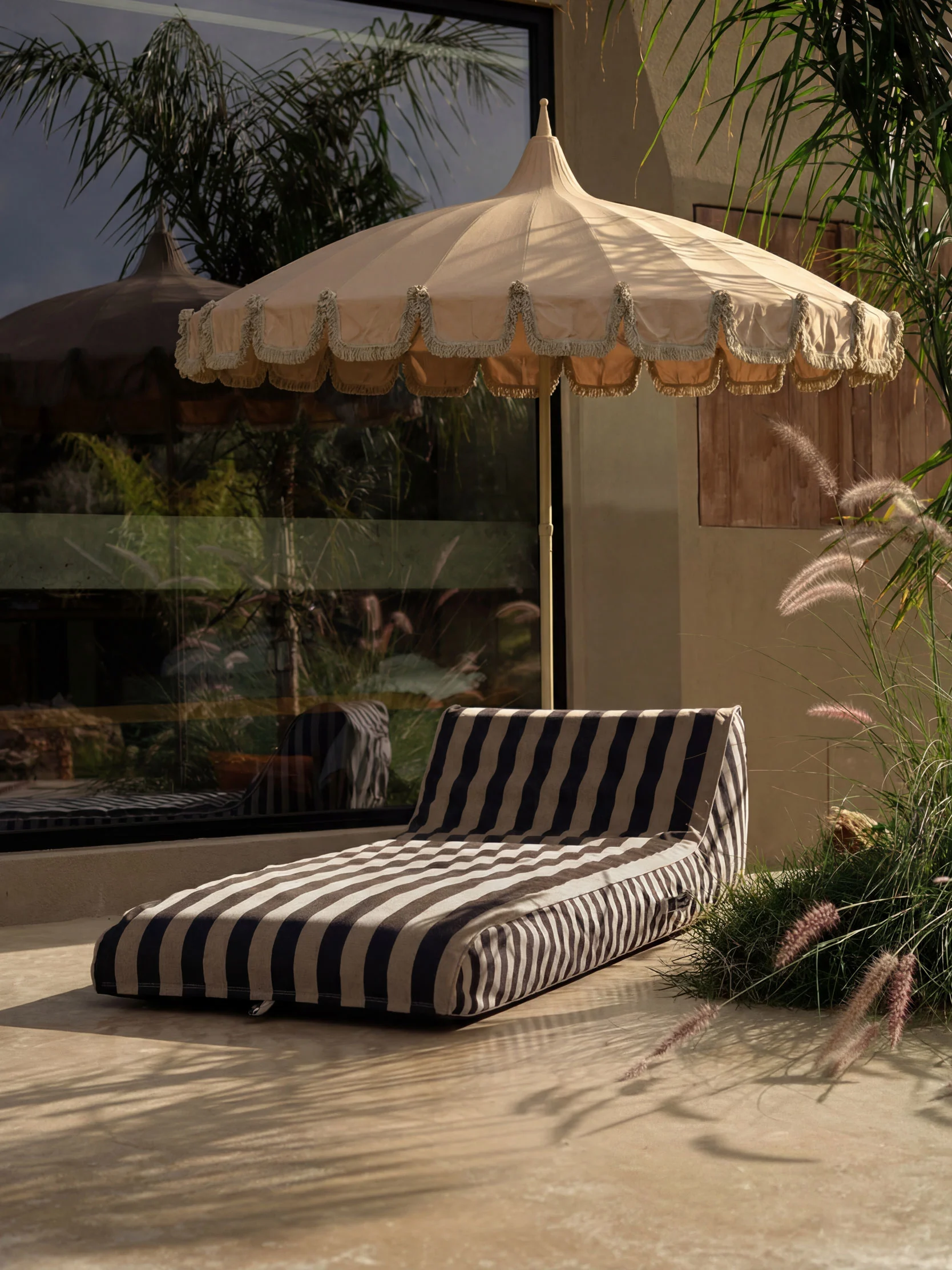 Patio extérieur avec parasol beige à franges et coussin de salon rayé sur sol clair, entouré de plantes.