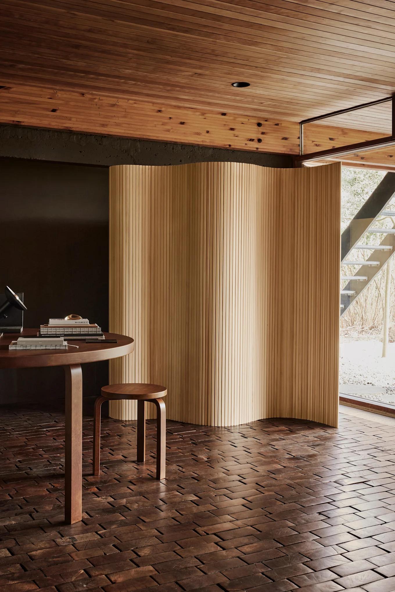 Pièce moderne avec une table ronde en bois, un petit tabouret et un écran en bois ondulé. Sol en blocs de bois foncé, plafond en lattes de bois clair et fenêtre donnant sur un escalier extérieur.