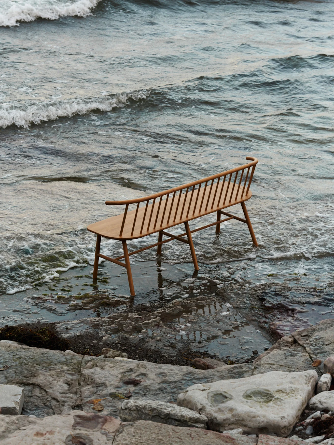Un banc en bois clair à lattes avec un dossier incurvé se dresse dans des eaux peu profondes sur une plage rocheuse avec de douces vagues.