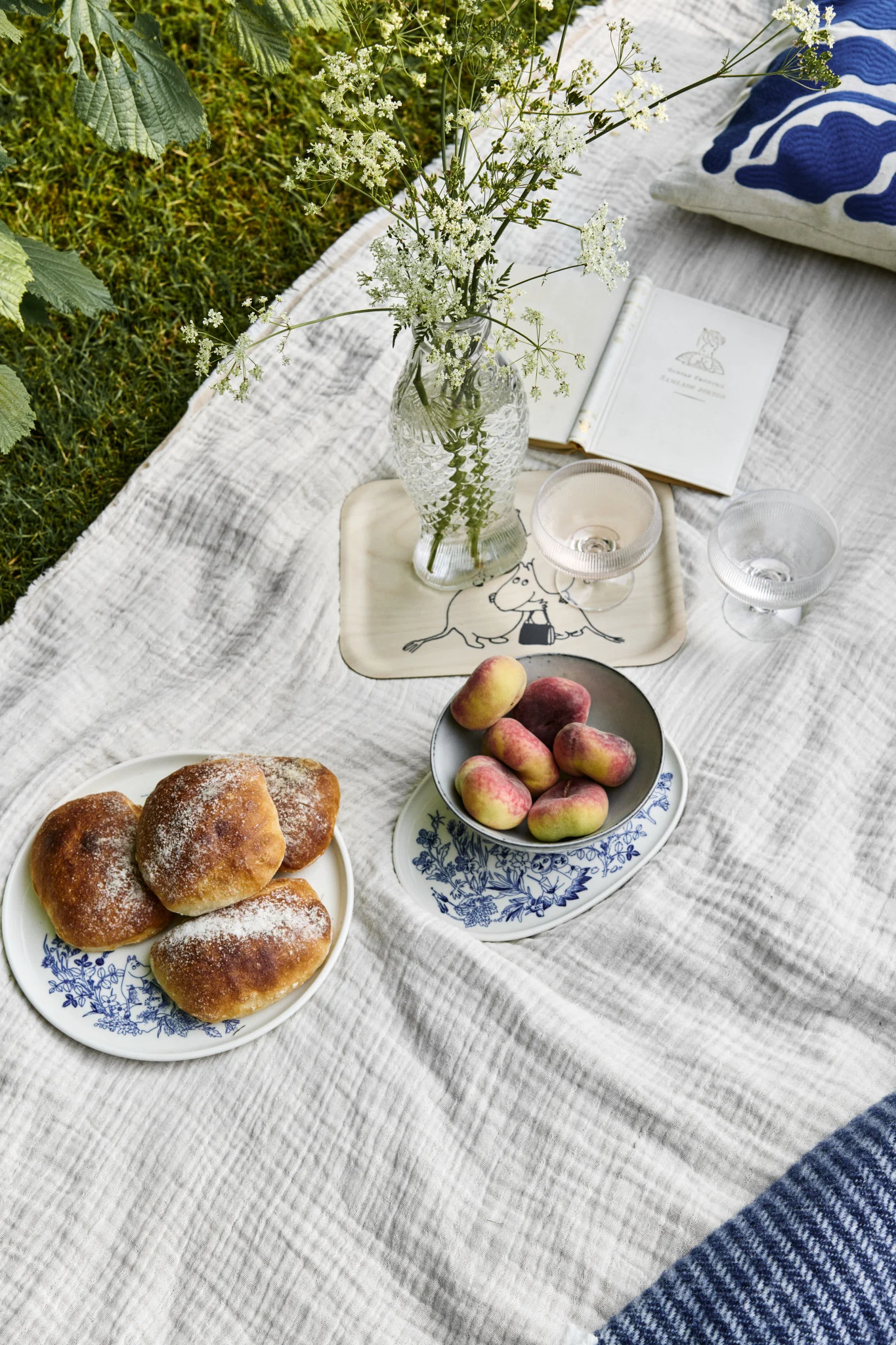 Pique-nique sur l'herbe avec des petits pains, des pêches plates, des fleurs sauvages dans un vase et un livre ouvert sur une nappe blanche.
