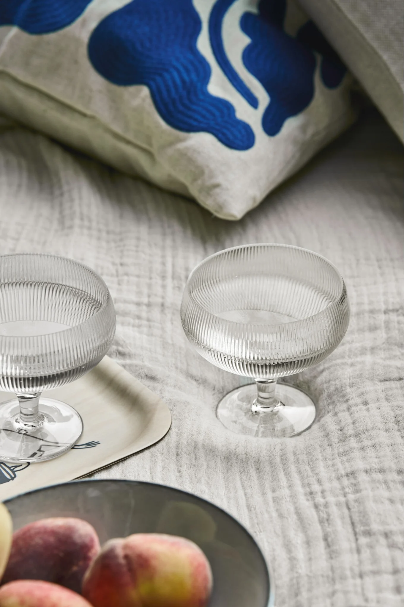 Deux coupes en verre nervurées, un bol de pêches et un coussin à motifs sur un textile clair.