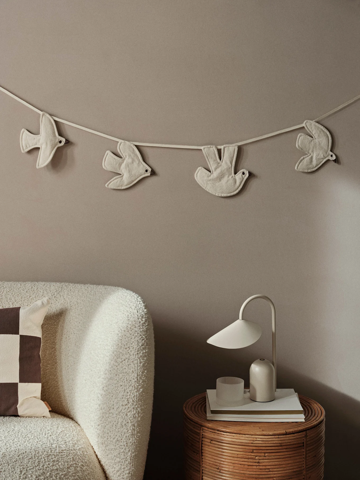 Guirlande d'oiseaux en tissu sur un mur neutre, au-dessus d'un canapé texturé et d'une table en rotin avec une lampe blanche.