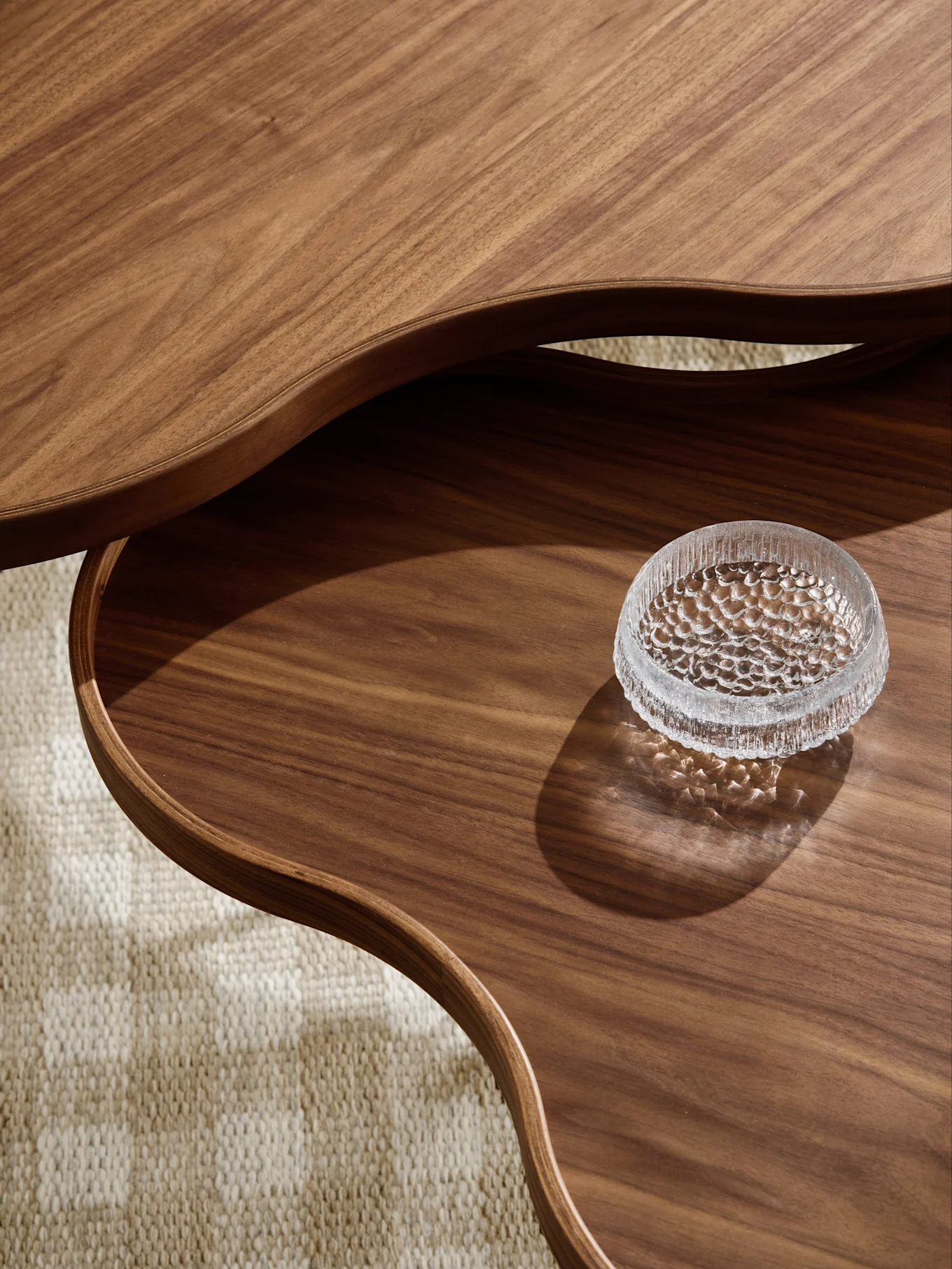 Gros plan d'une table basse en bois à plusieurs niveaux, bords ondulés, bol en verre texturé sur le niveau inférieur, sur tapis.