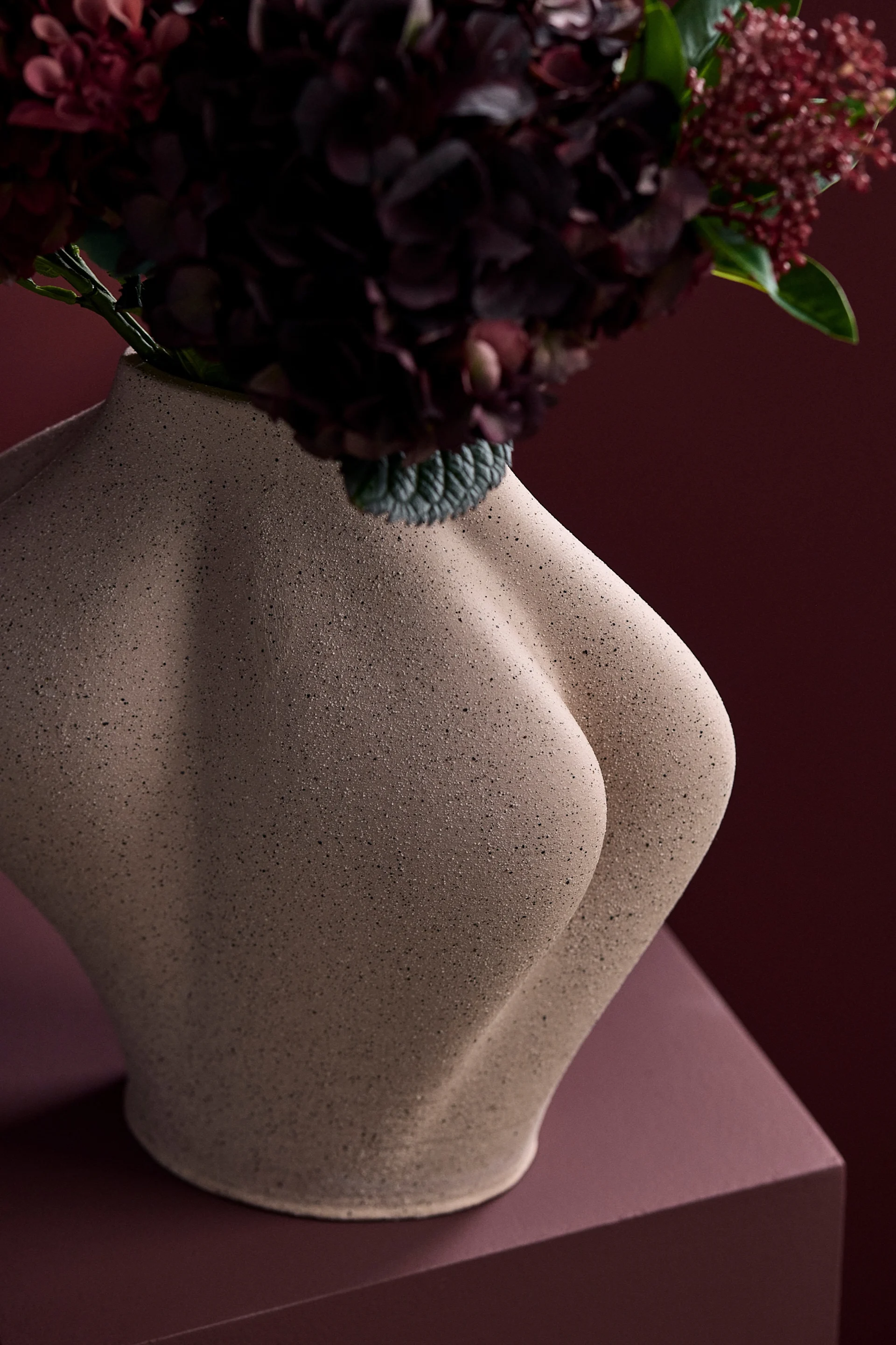 Vase sculptural de couleur beige, en forme de torse humain, contenant des fleurs rouge foncé sur un fond rouge assorti.