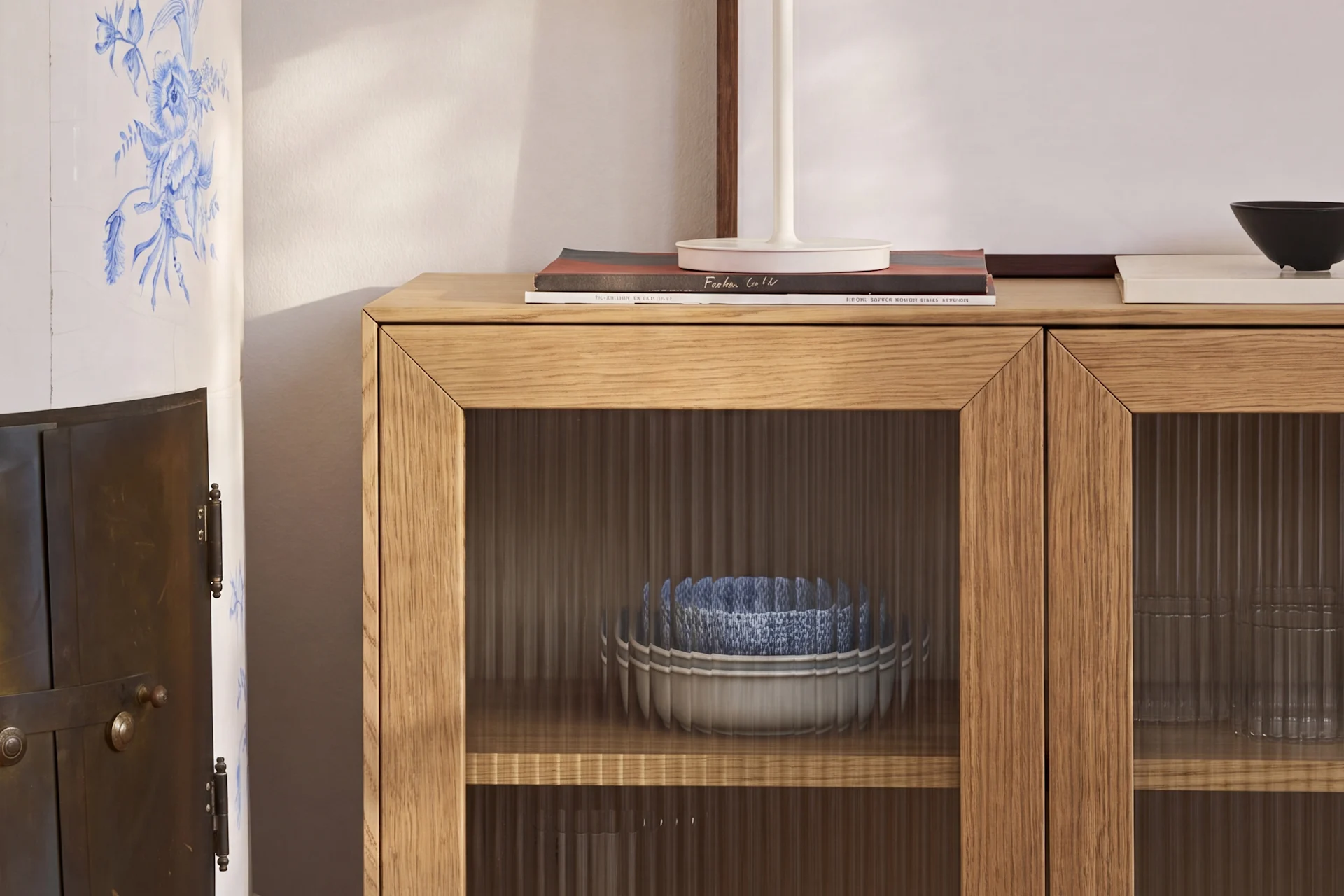 Un meuble en bois clair avec des portes en verre cannelé contient des bols bleus et blancs, à côté d'un poêle en céramique à motifs floraux bleus.