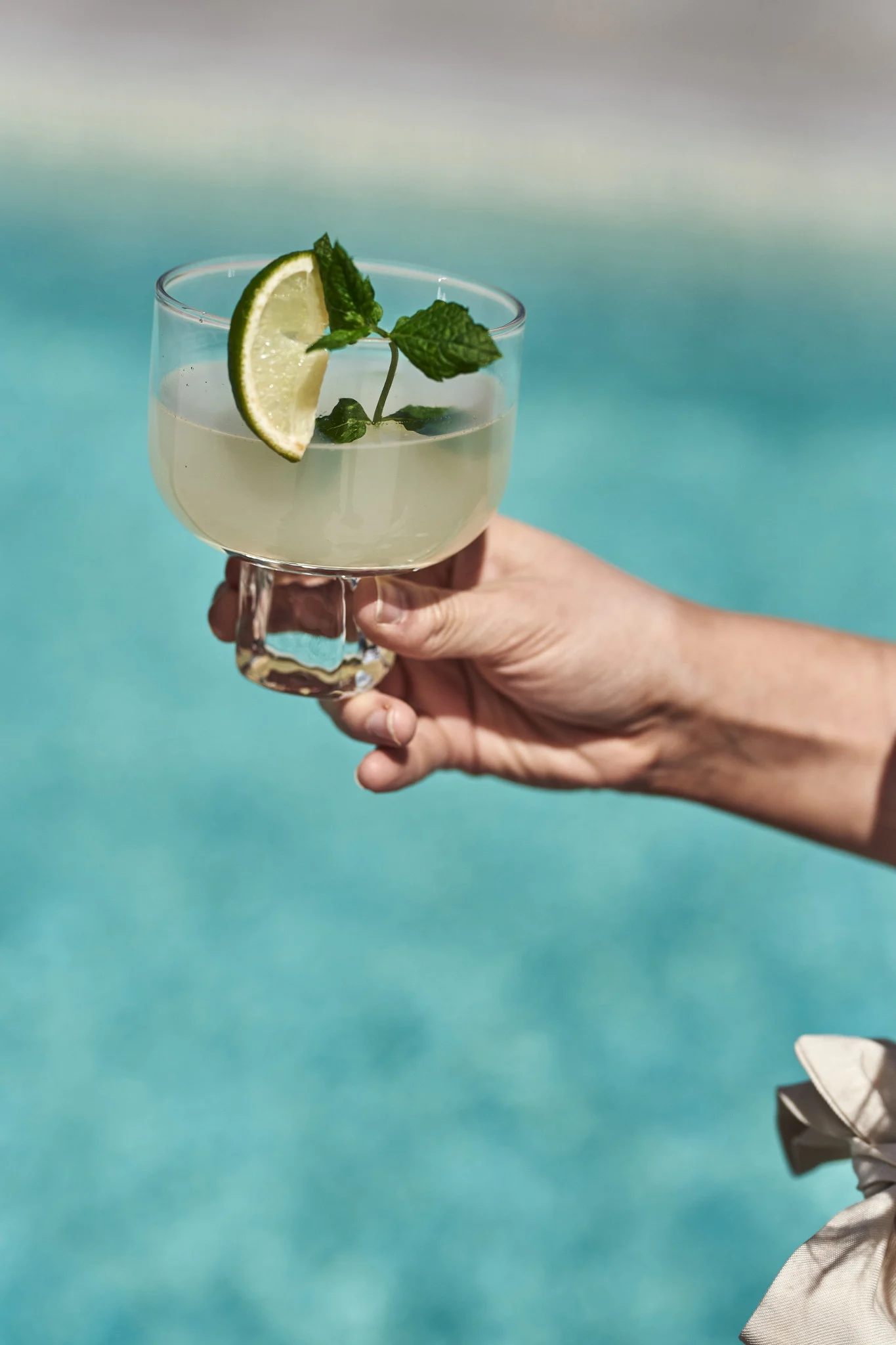 Découvrez ces cocktails et mocktails d'été simples et rafraîchissants. Ici, une main tient un mojito avec un quartier de citron vert au bord d'une piscine.